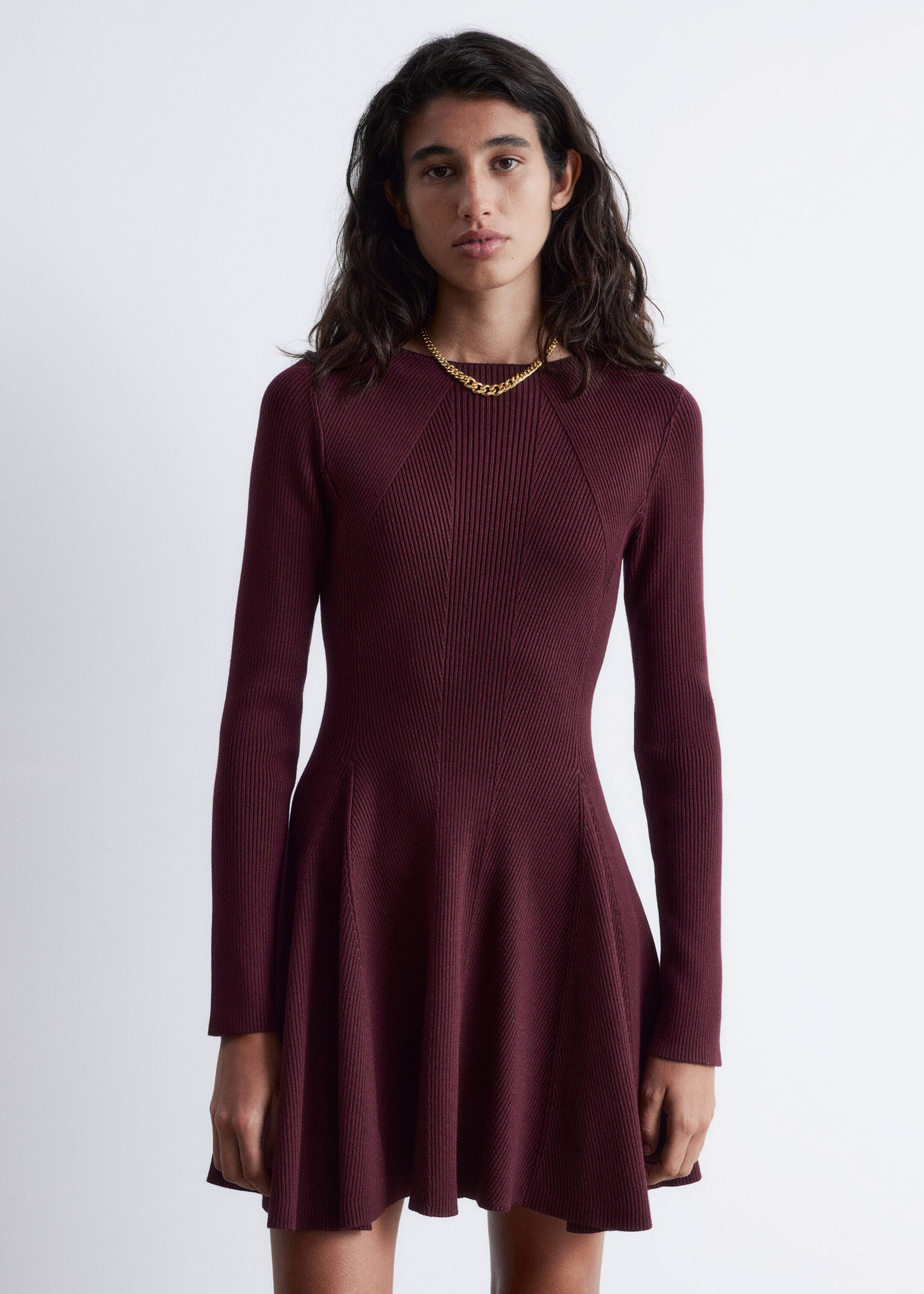 Agrandir l'image: Une robe mini en tricot côtelé rouge profond, avec un col montant, des manches longues, une taille ajustée et une jupe évasée à panneaux, est portée par un mannequin. Un collier chaîne de ton doré est porté par-dessus la robe.