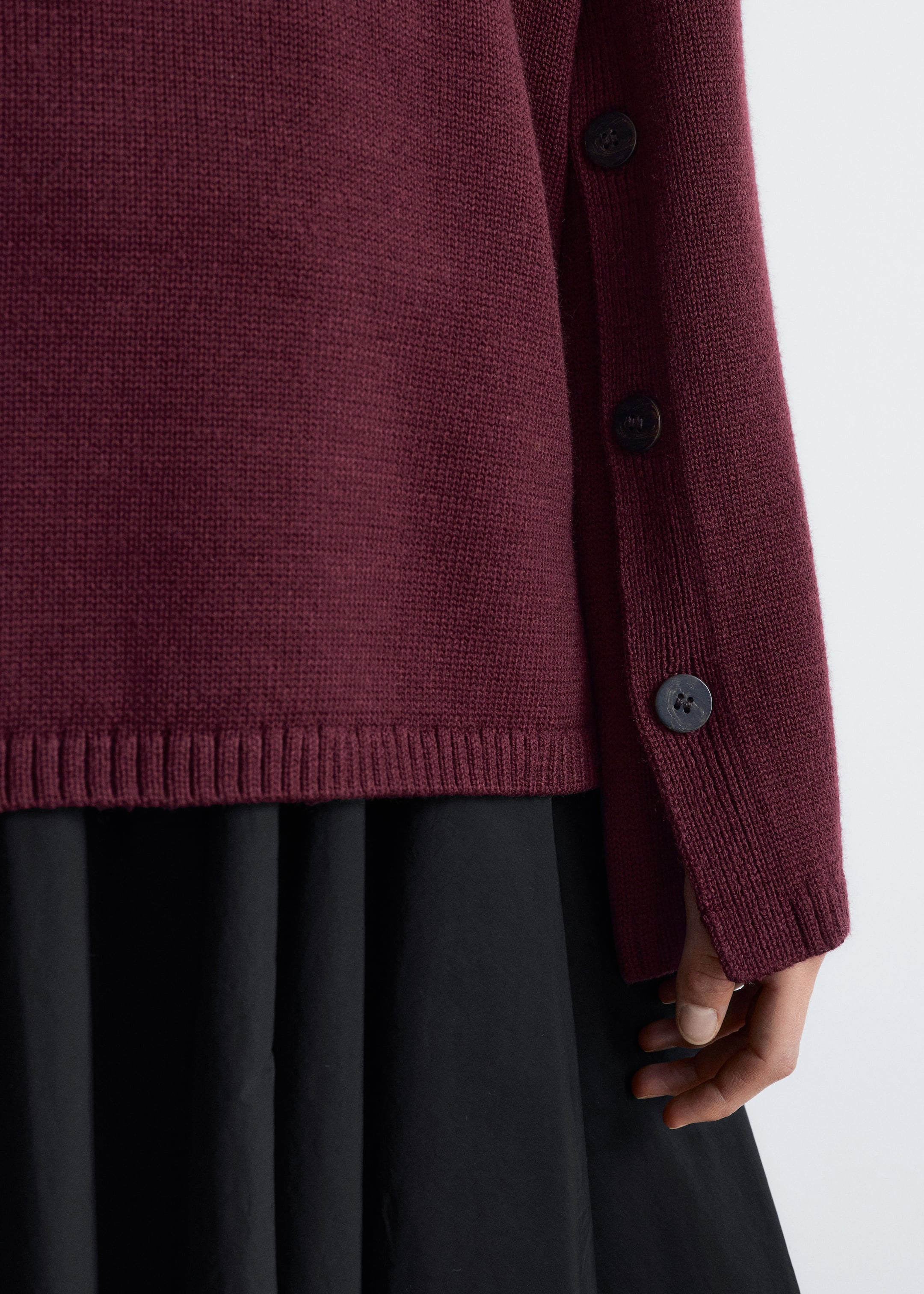 View larger image: A burgundy knit sweater is showing a ribbed hem and a vertical line of dark, round buttons along a side opening, extending down the length of the sleeve. The sweater is layered over a black flowing garment, with a hand visible near the sleeve's ribbed cuff.