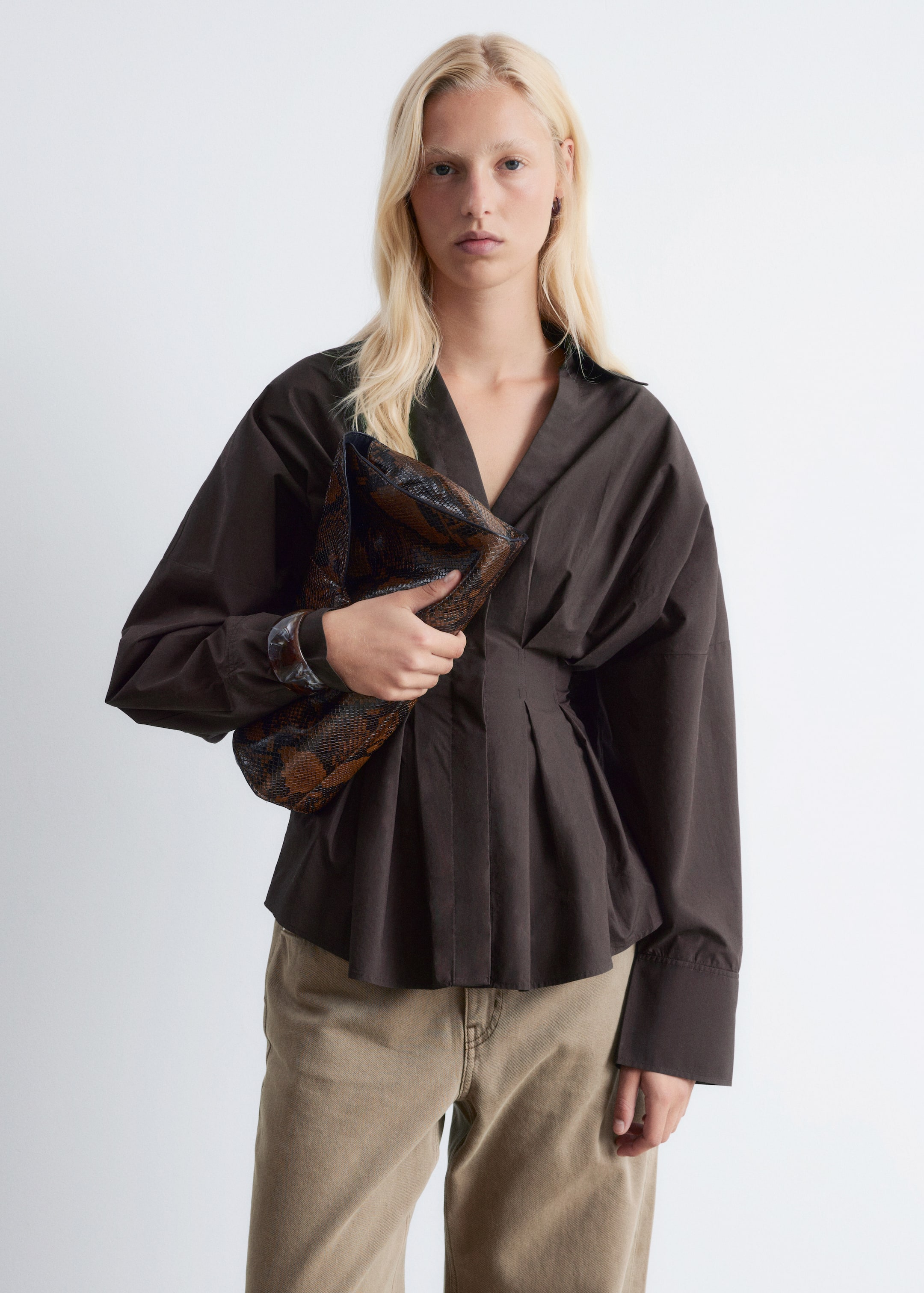 Agrandir l'image: Une chemise marron foncé à manches longues est portée, avec un col en V ouvert et des poignets larges boutonnés. La chemise est cintrée à la taille, d'où des plis s'évasent vers le bas, créant un effet péplum.