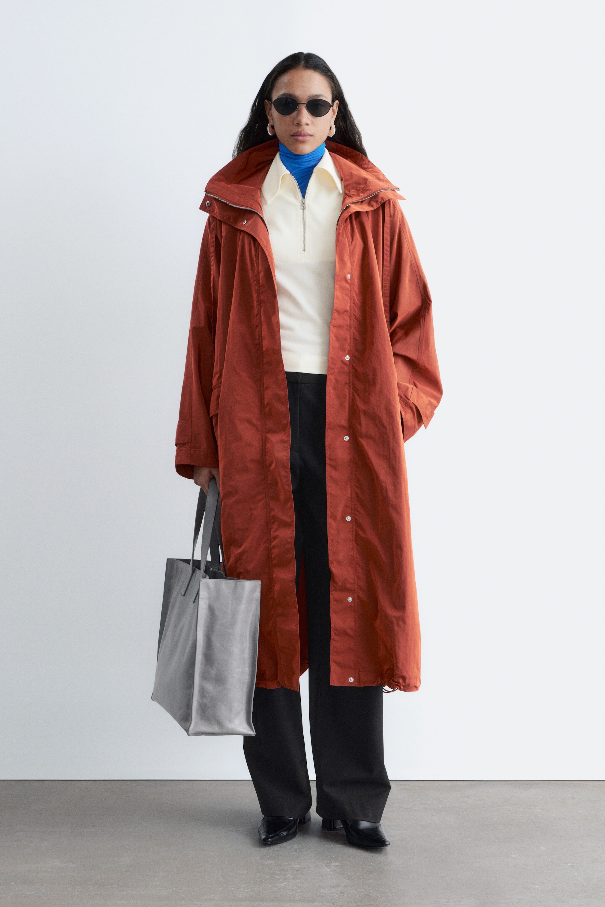 Agrandir l'image: Un long manteau couleur rouille présente une coupe droite et décontractée, s'étendant au-delà des genoux. Il est doté d'un col montant avec une capuche froncée et de fermetures à boutons-pression sur le devant, superposé à un polo crème à quart de zip et un col roulé bleu.