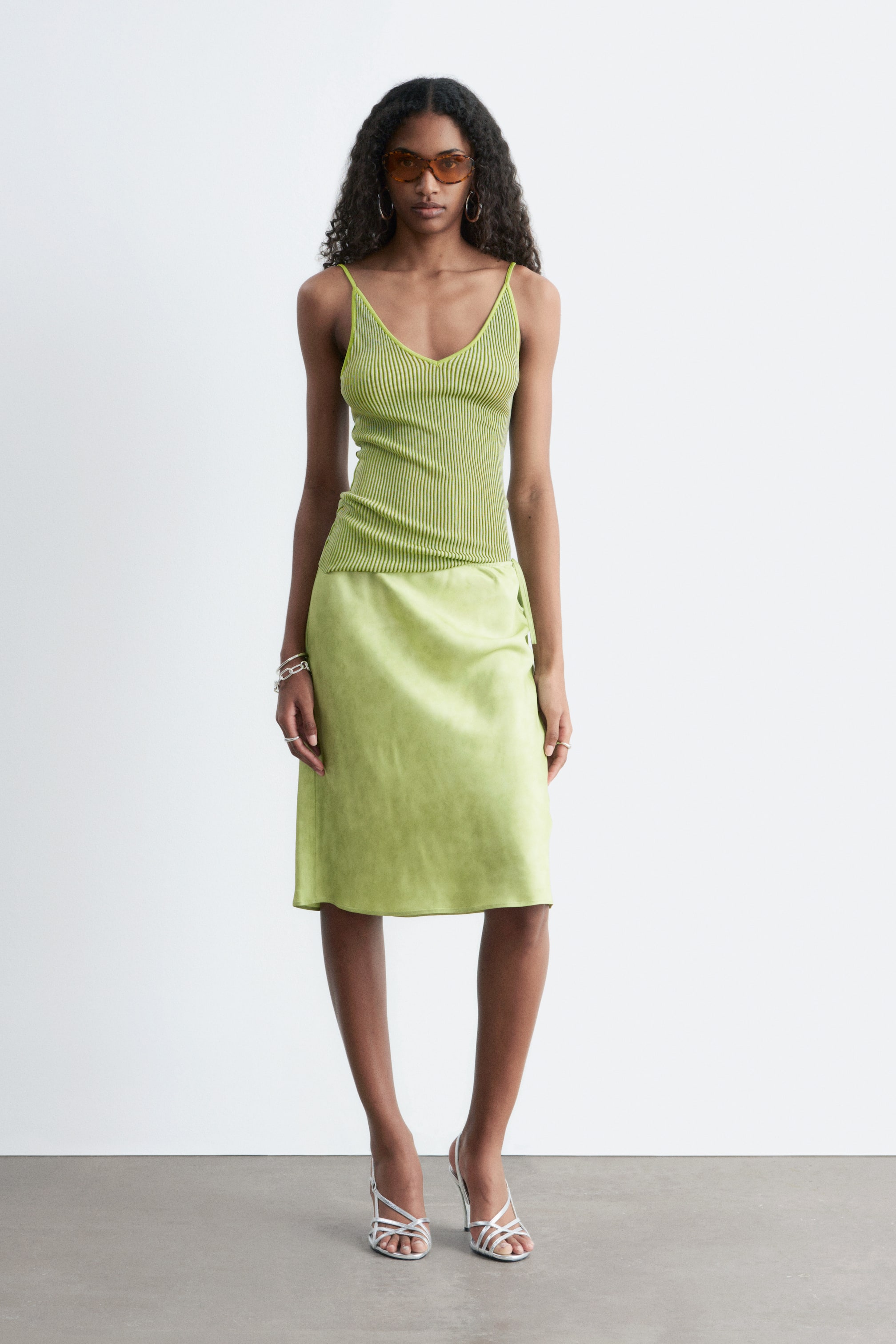 Ingrandisci l'immagine: Un completo verde lime a due pezzi viene mostrato dal davanti. Il top a canotta aderente in maglia a coste presenta spalline sottili, uno scollo a V e sottili righe orizzontali scure, mentre la gonna coordinata in raso cade a lunghezza midi con un delicato dettaglio di nodo laterale sul fianco.