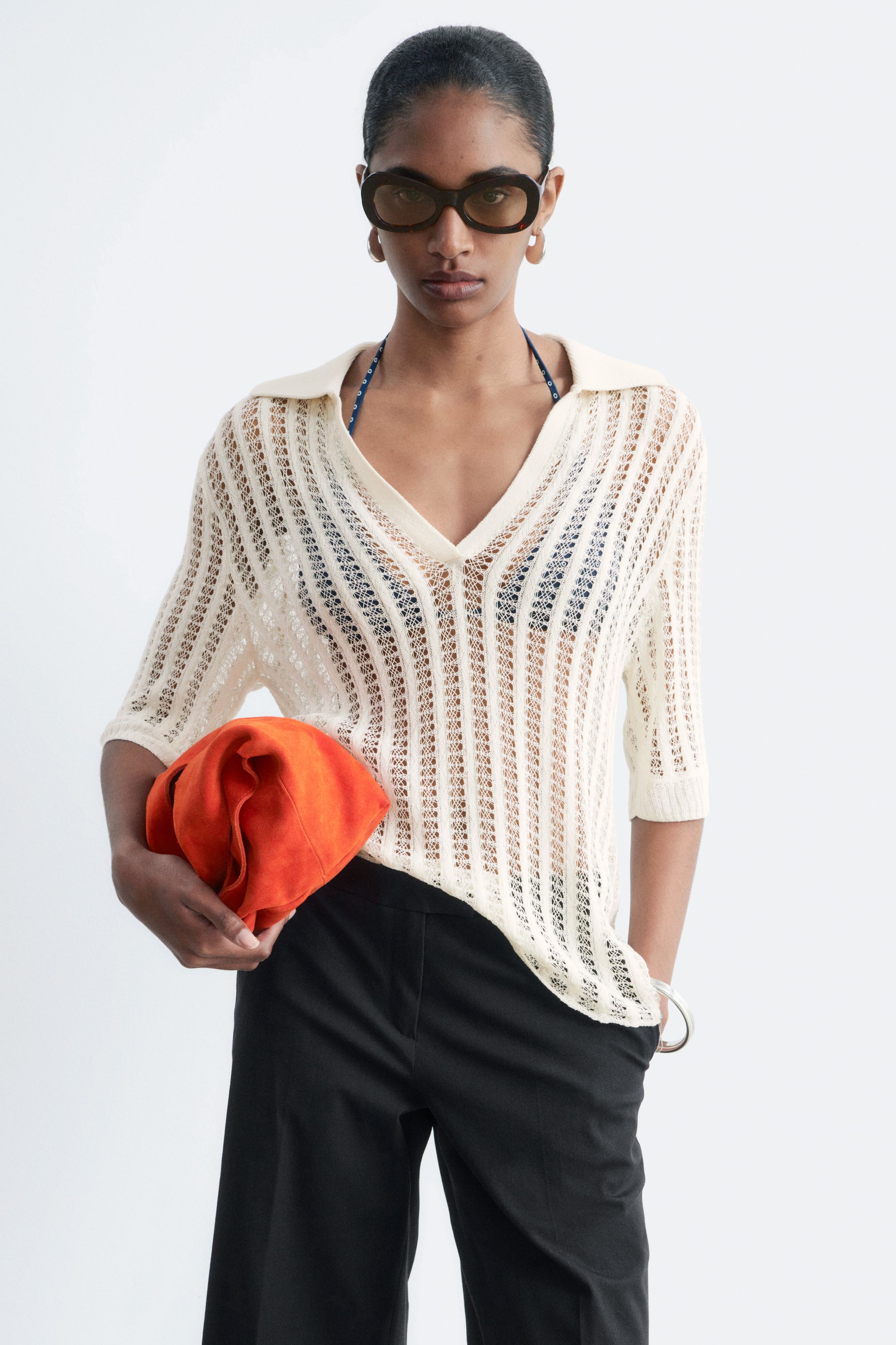 Grösseres Bild ansehen: Ein cremefarbenes, offen gestricktes Poloshirt mit V-Ausschnitt, Kragen und gerippten Bündchen wird vom Model getragen. Darunter ist ein blau gemustertes Bikinioberteil zu sehen, und das Model hält eine orangefarbene Clutch-Tasche.