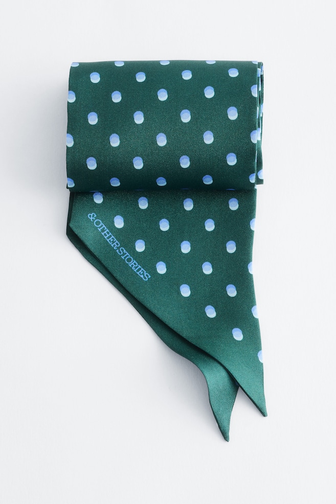Ein schmaler, dunkelgrüner Seidenschal mit einem durchgehenden Muster aus hellblauen Polka Dots ist gefaltet dargestellt, wobei die schrägen, spitz zulaufenden Enden nach unten hängen. Der Markenname â& OTHER STORIESâ ist in Hellblau diagonal über eine der spitzen Enden gedruckt. - 1