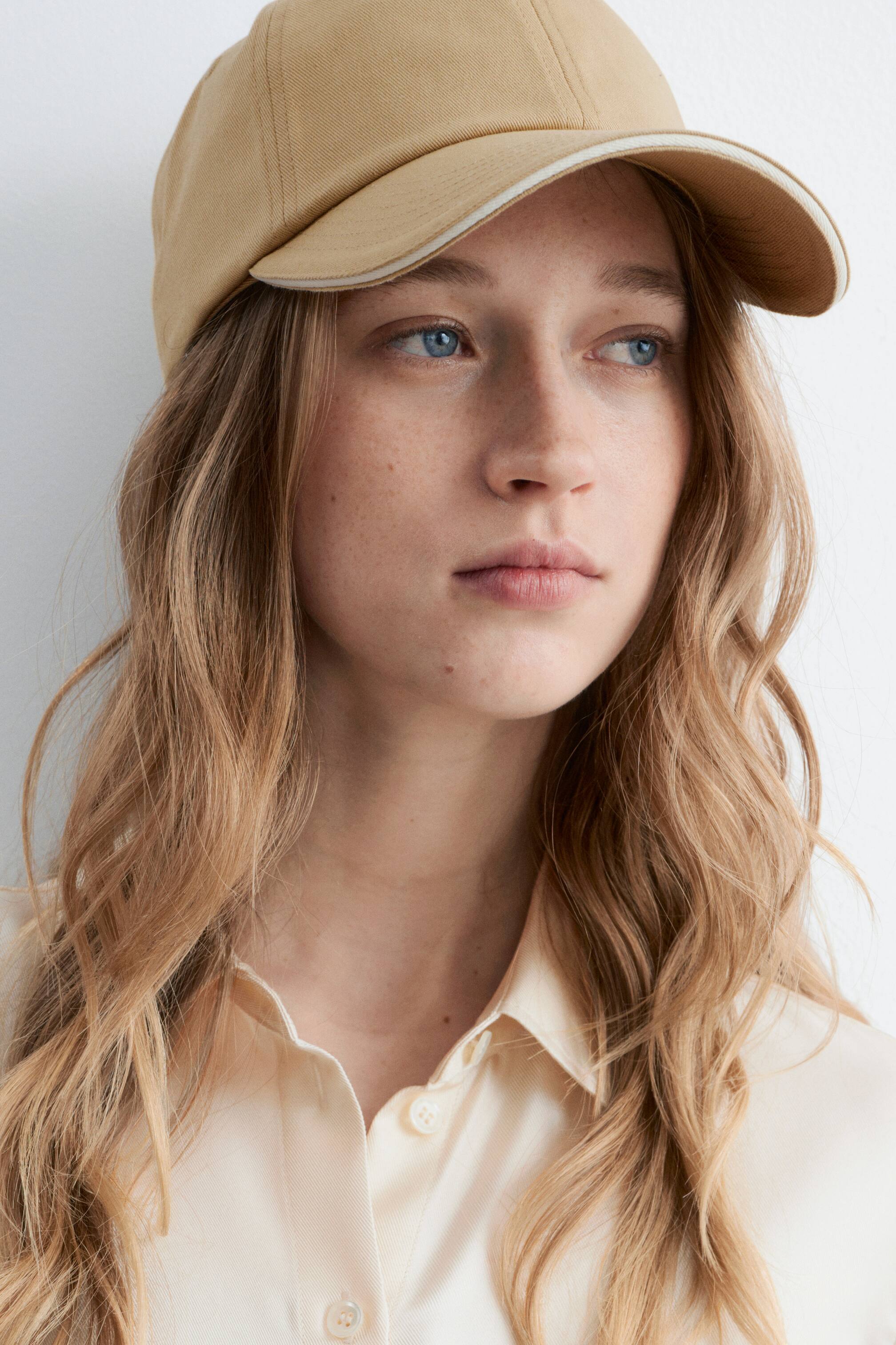 Ingrandisci l'immagine: Un cappellino da baseball beige con un design a pannelli, una visiera curva con un bordo bianco a contrasto e cuciture visibili, indossato dal modello.