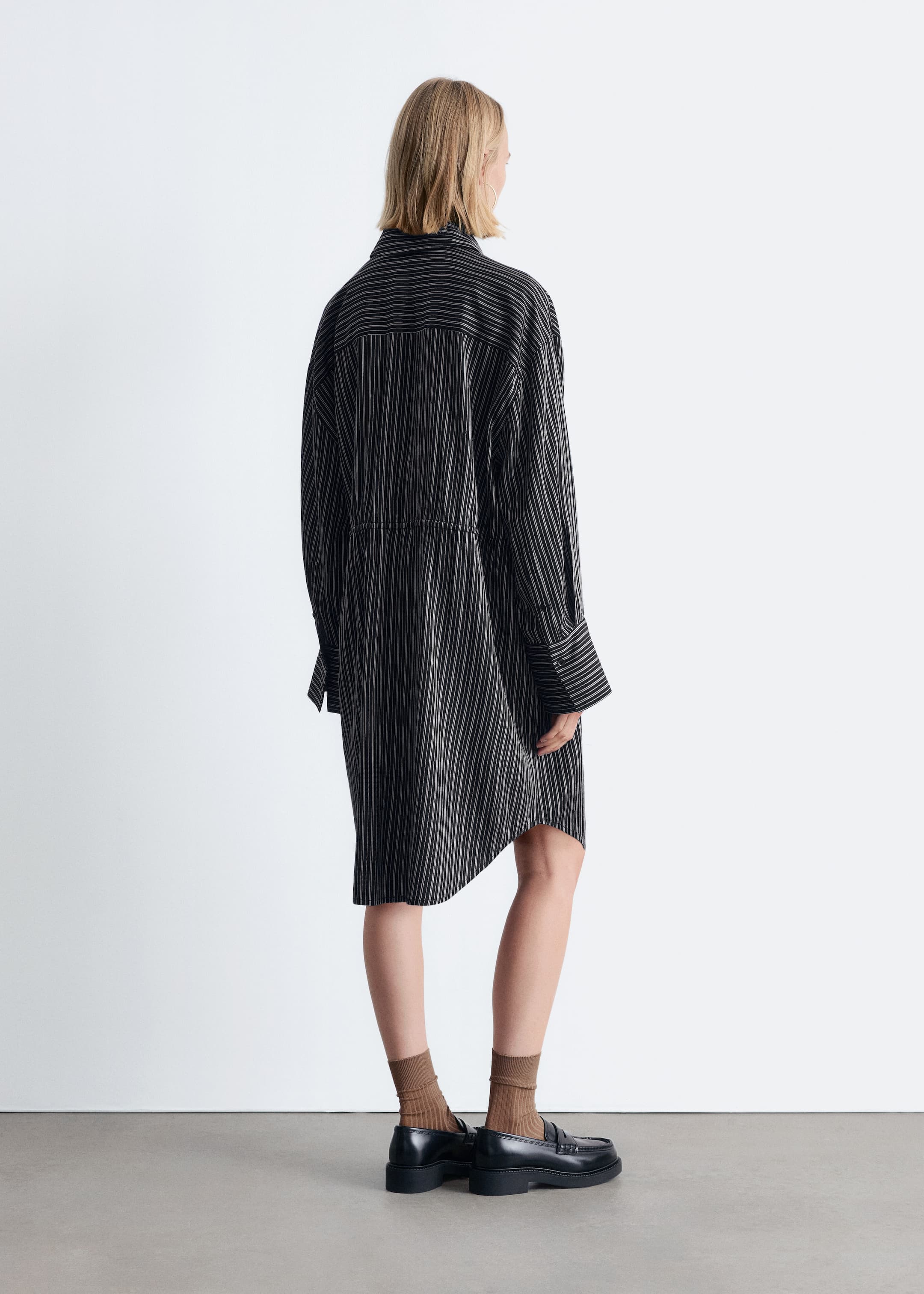 Agrandir l'image: Une robe-chemise noire surdimensionnée avec de fines rayures verticales blanches est montrée de dos. Elle présente un col classique, des poignets larges boutonnés, un détail cintré à la taille arrière et un ourlet asymétrique qui monte plus haut d'un côté.