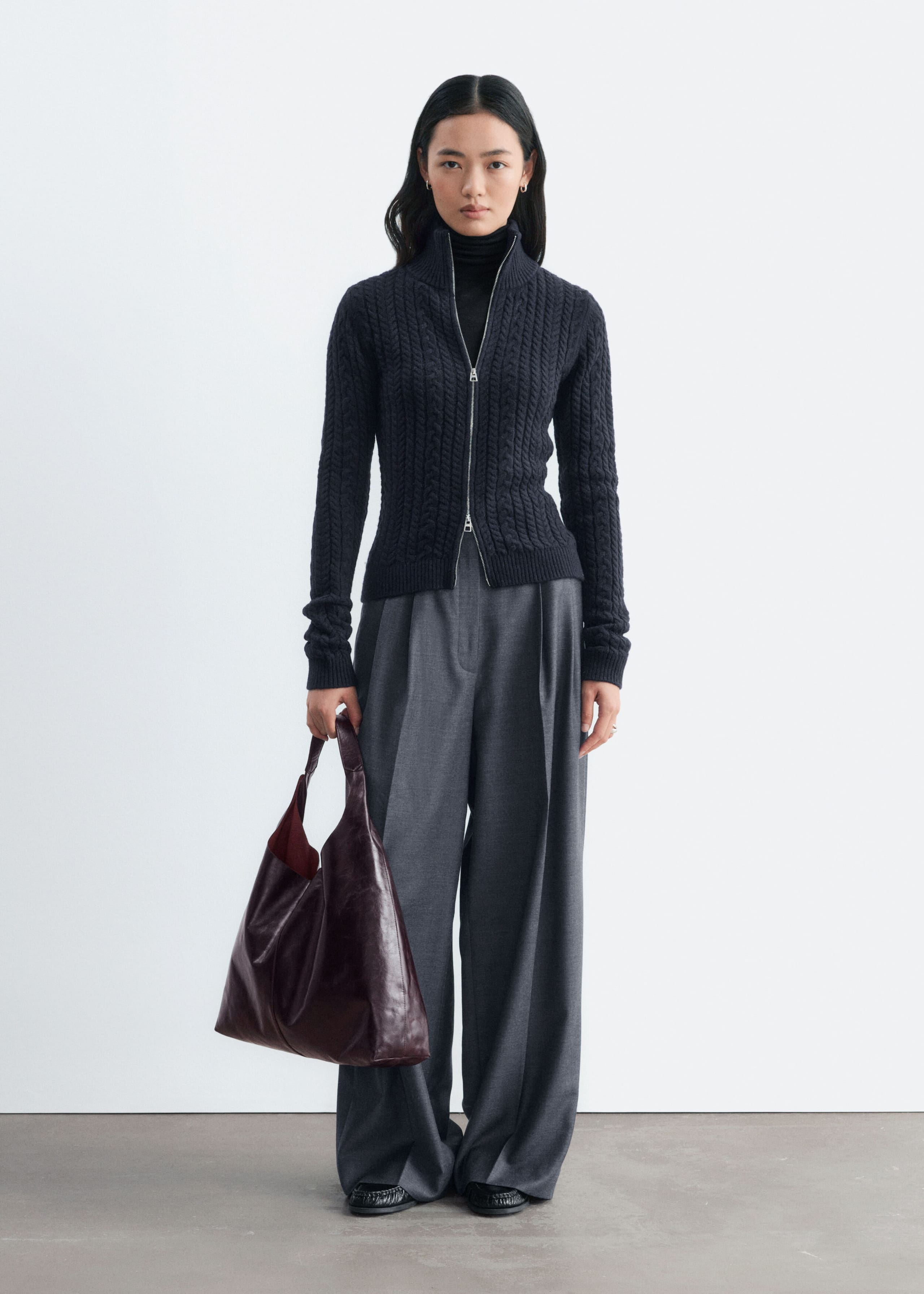 The model is wearing a dark grey cable-knit cardigan, featuring a slim fit, a high collar, and a partially open two-way front zip. The cardigan is layered over a black roll-neck and styled with grey wide-leg trousers.
