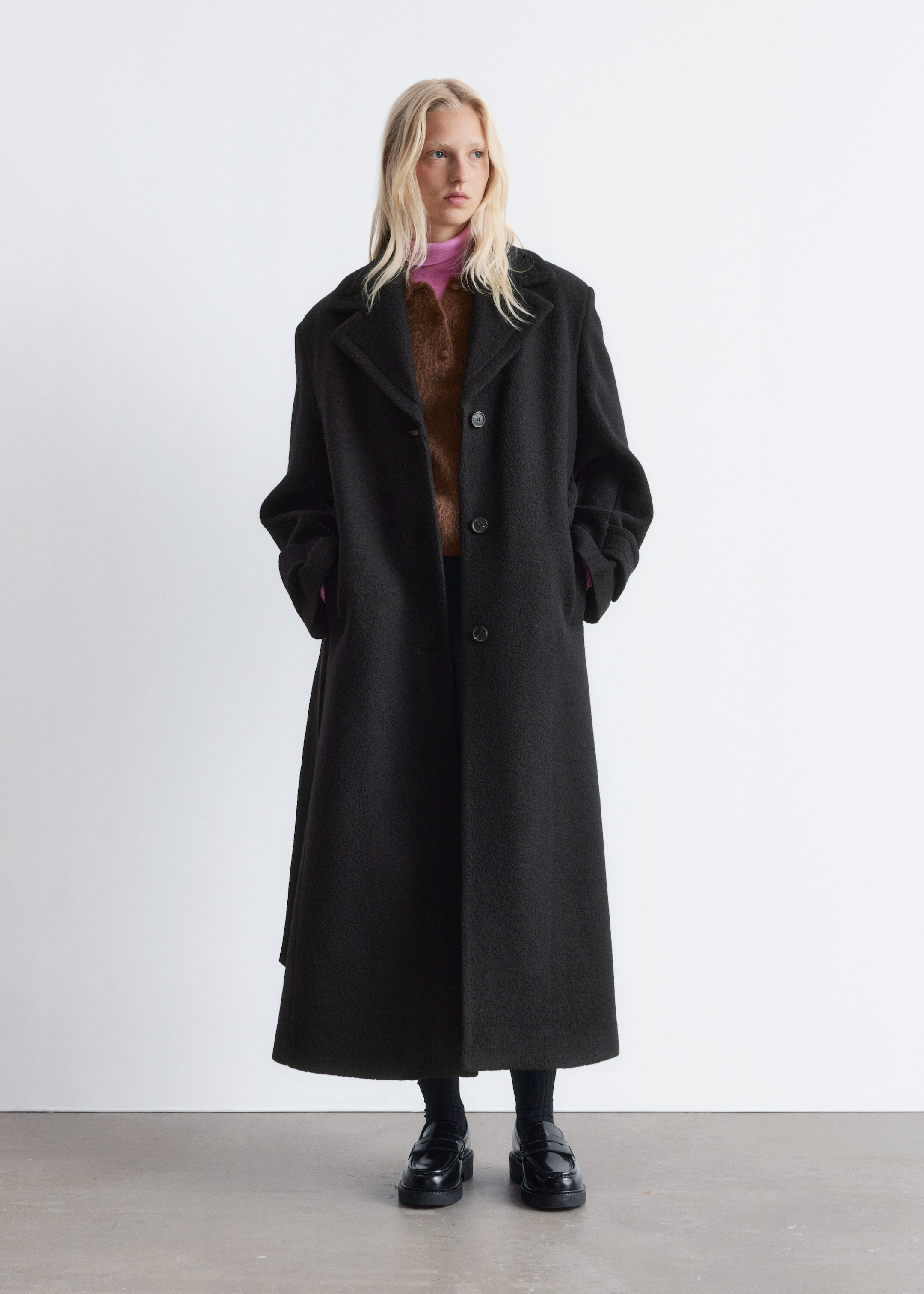 Agrandir l'image: Un long manteau noir est présenté de face, avec de larges revers crantés et une fermeture à deux boutons. Le manteau est conçu avec une coupe décontractée et ample, les mains du porteur placées dans les poches latérales visibles.