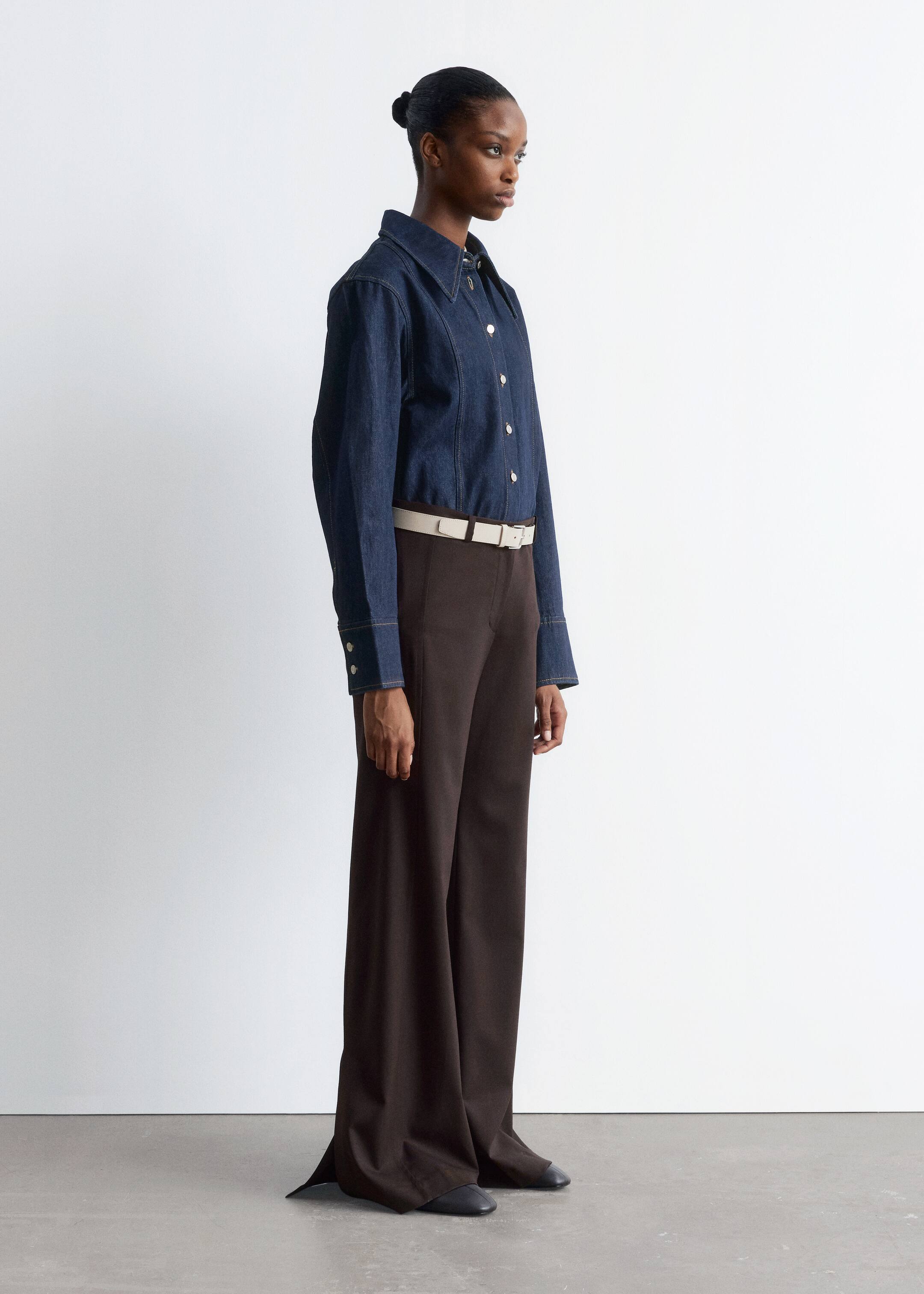 Ingrandisci l'immagine: Una camicia di jeans blu scuro con colletto a punta, chiusure con bottoni color argento e cuciture a contrasto è indossata dal modello. La camicia è abbinata a pantaloni scuri e una cintura chiara.