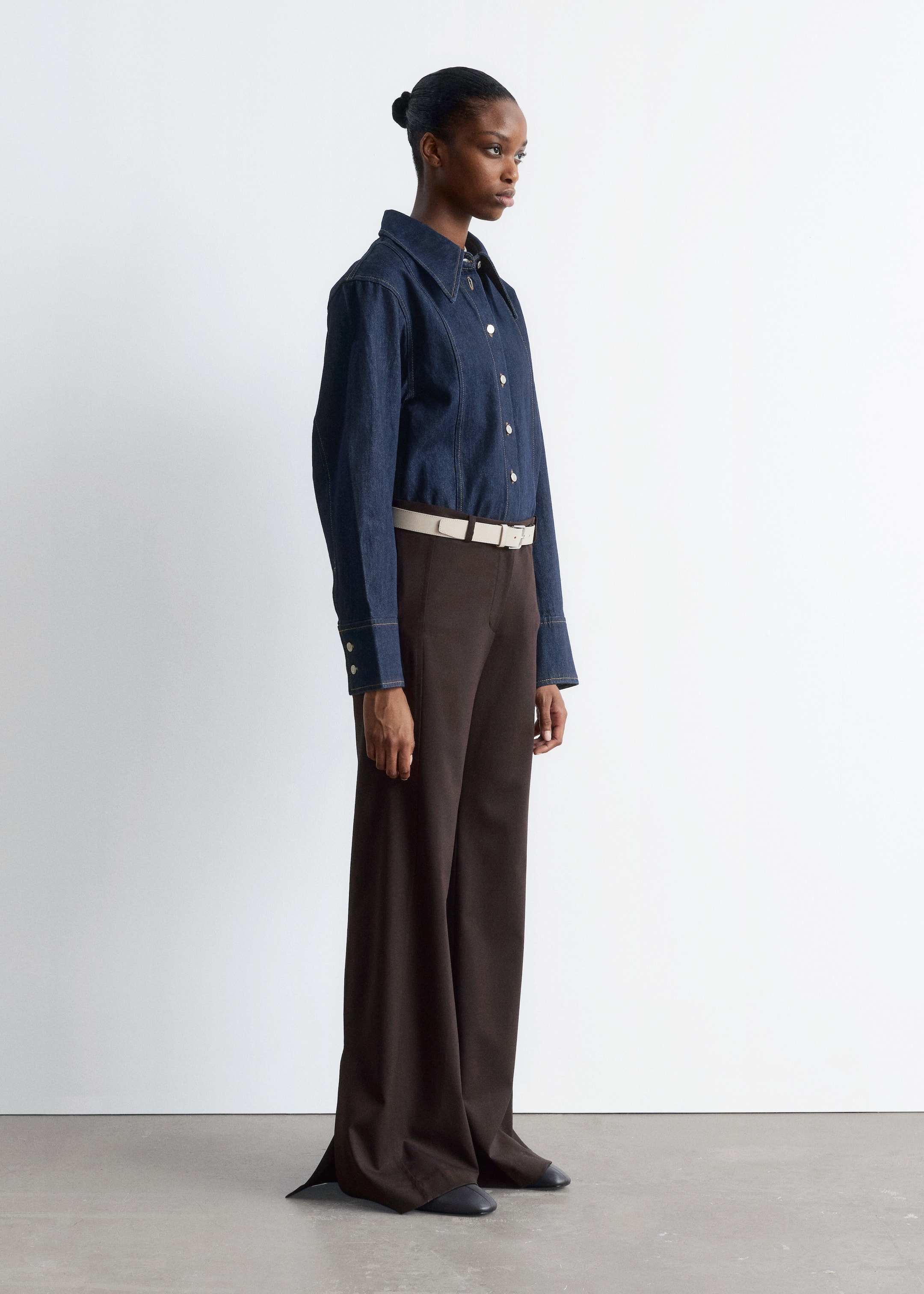 Agrandir l'image: Une chemise en denim bleu foncé avec un col pointu, des fermetures à boutons de couleur argentée et des surpiqûres contrastées est portée par le mannequin. La chemise est assortie à un pantalon foncé et une ceinture claire.