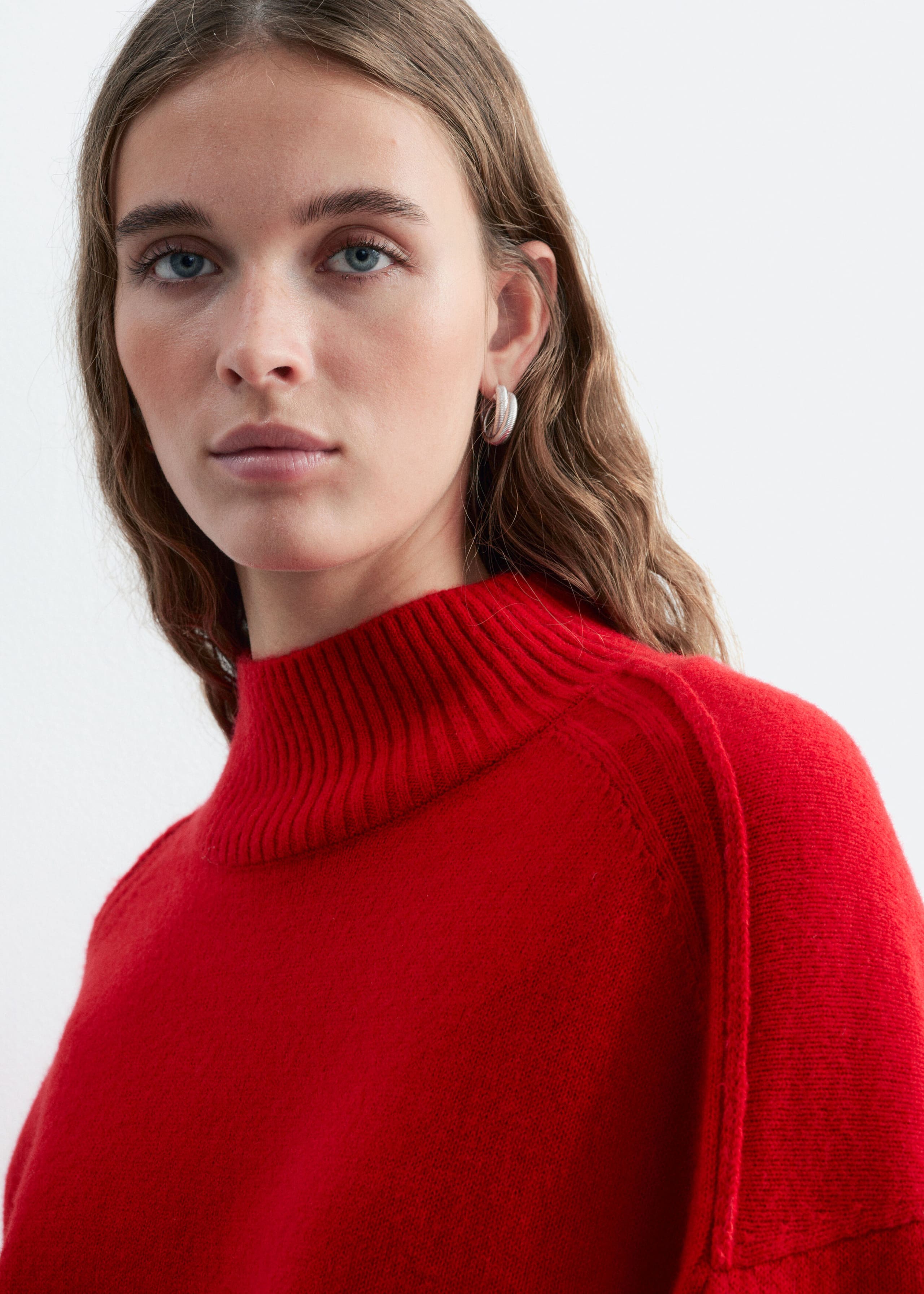 This red mock neck sweater, featuring a ribbed collar and forward shoulder seams, is being worn by the model. The close-up shot highlights the soft texture of the knit fabric.
