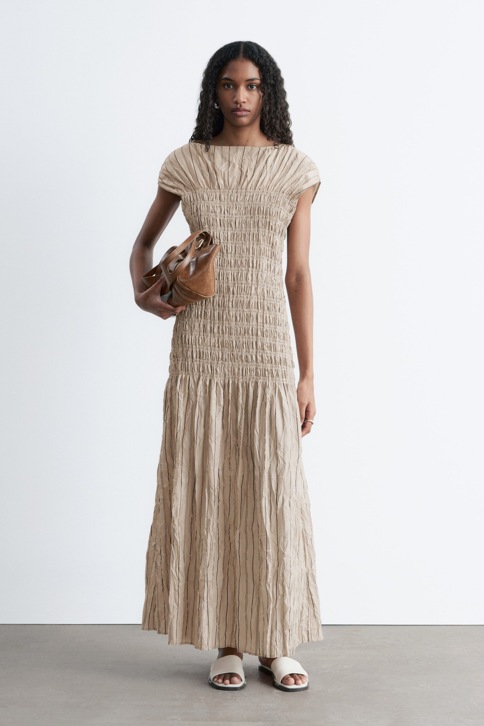 Ver imagem maior: Um vestido maxi em bege claro apresenta um decote barco e mangas curtas, com um corpete extensivamente franzido que cria uma aparência ajustada e texturizada. O vestido flui para uma saia em linha A com texturas enrugadas verticais que formam linhas subtis mais escuras.
