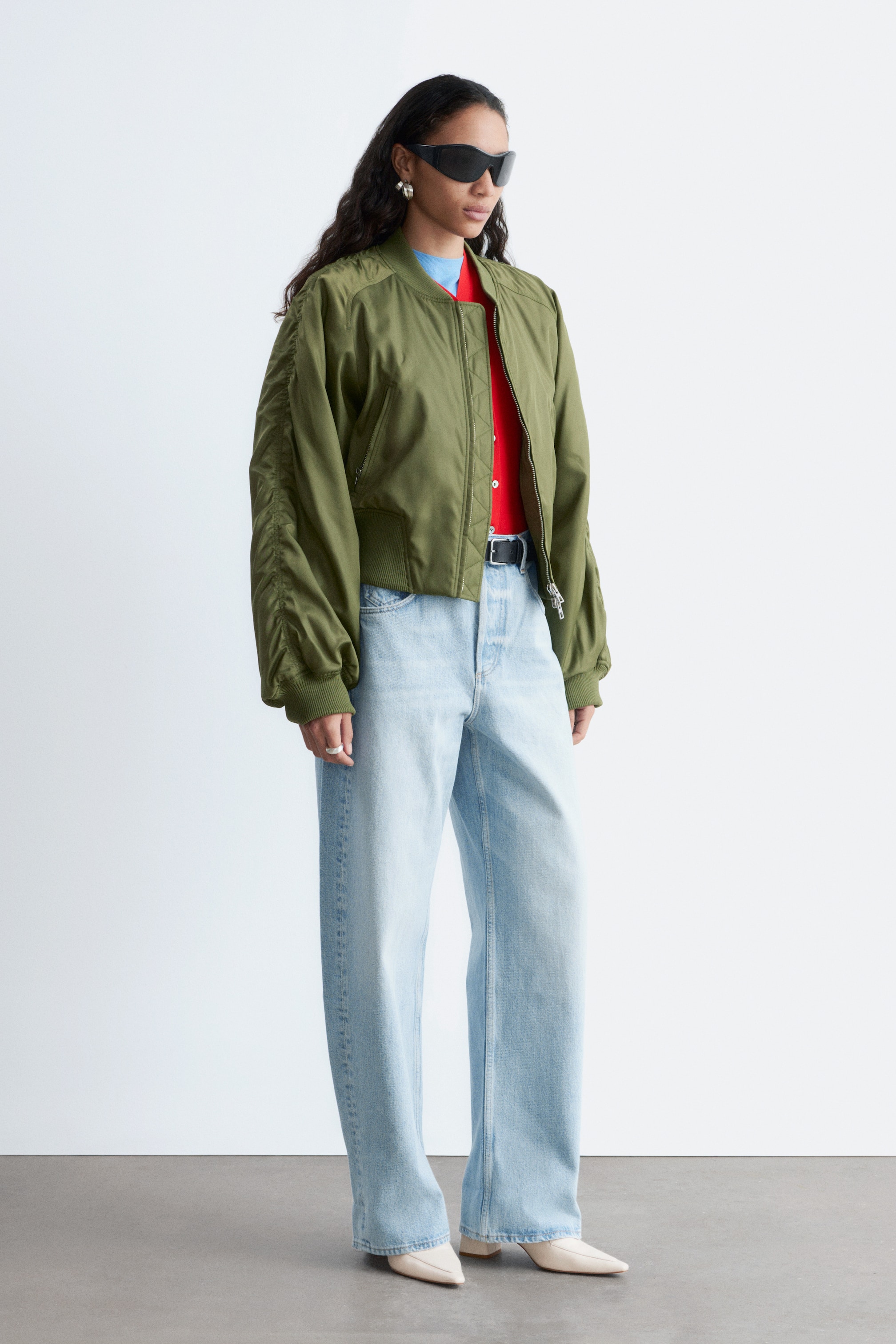 Se større billede: En olivengrøn bomberjakke præsenteres, med en ribbet stand-up krave, ribbede manchetter og en ribbet kant. Dens frontlynlås er åben, hvilket afslører et kontrasterende indre lag, og den inkluderer en lynlåslomme på venstre ærme med rynkedetaljer langs de ydre sømme.