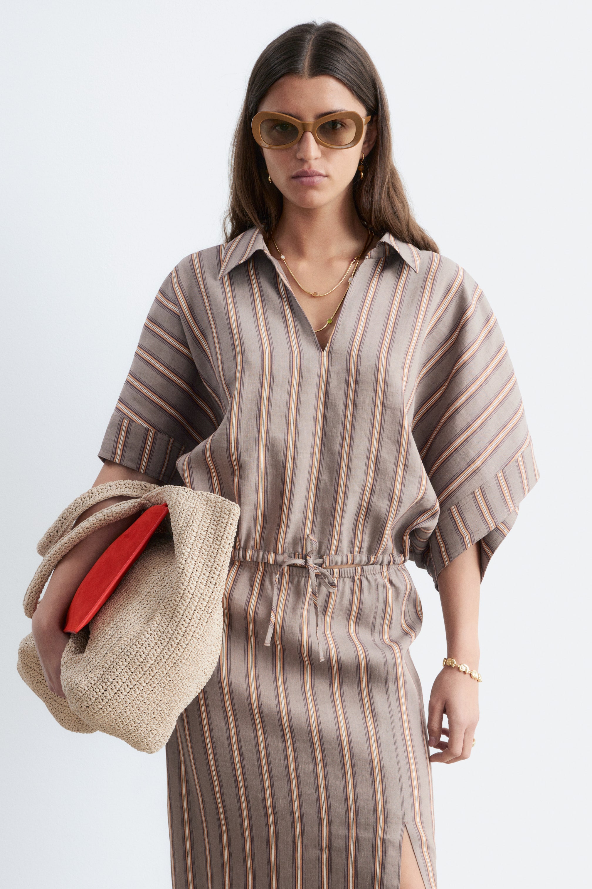 Ver imagen más grande: Un vestido midi a rayas presenta una base de color gris marrón con rayas verticales en naranja y crema, un cuello en punta y una abertura en forma de V. Está diseñado con mangas cortas y anchas, cintura elástica con cordón ajustable, y una abertura lateral en la falda.