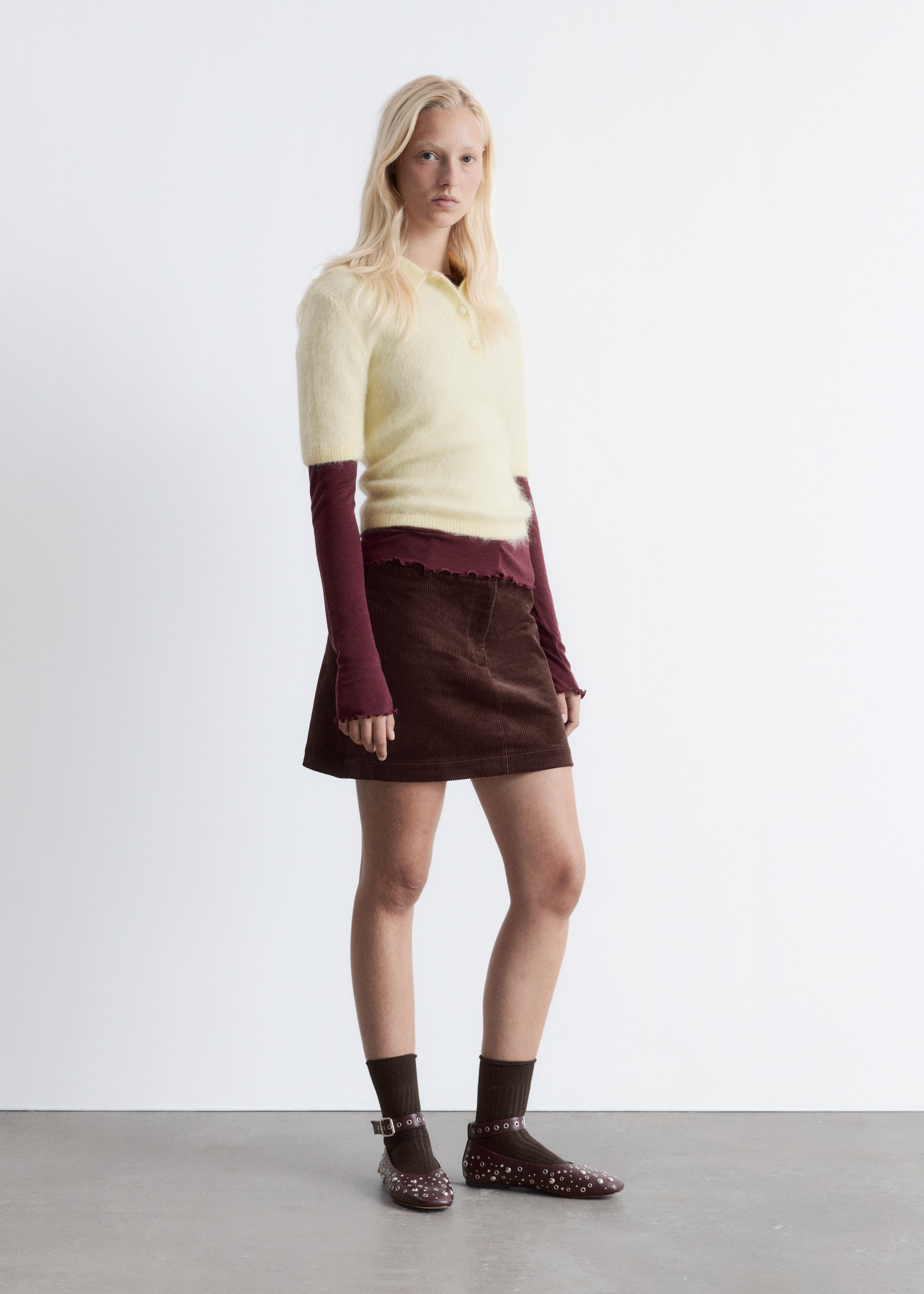 View larger image: A light yellow fuzzy short-sleeved polo shirt with a three-button placket is being layered over a dark red long-sleeved top, which features lettuce hems at the waist and cuffs. A brown corduroy A-line mini skirt with a lettuce hem is also being worn, along with dark brown ribbed ankle socks and dark brown Mary Jane flats adorned with multiple silver eyelets and buckled straps.