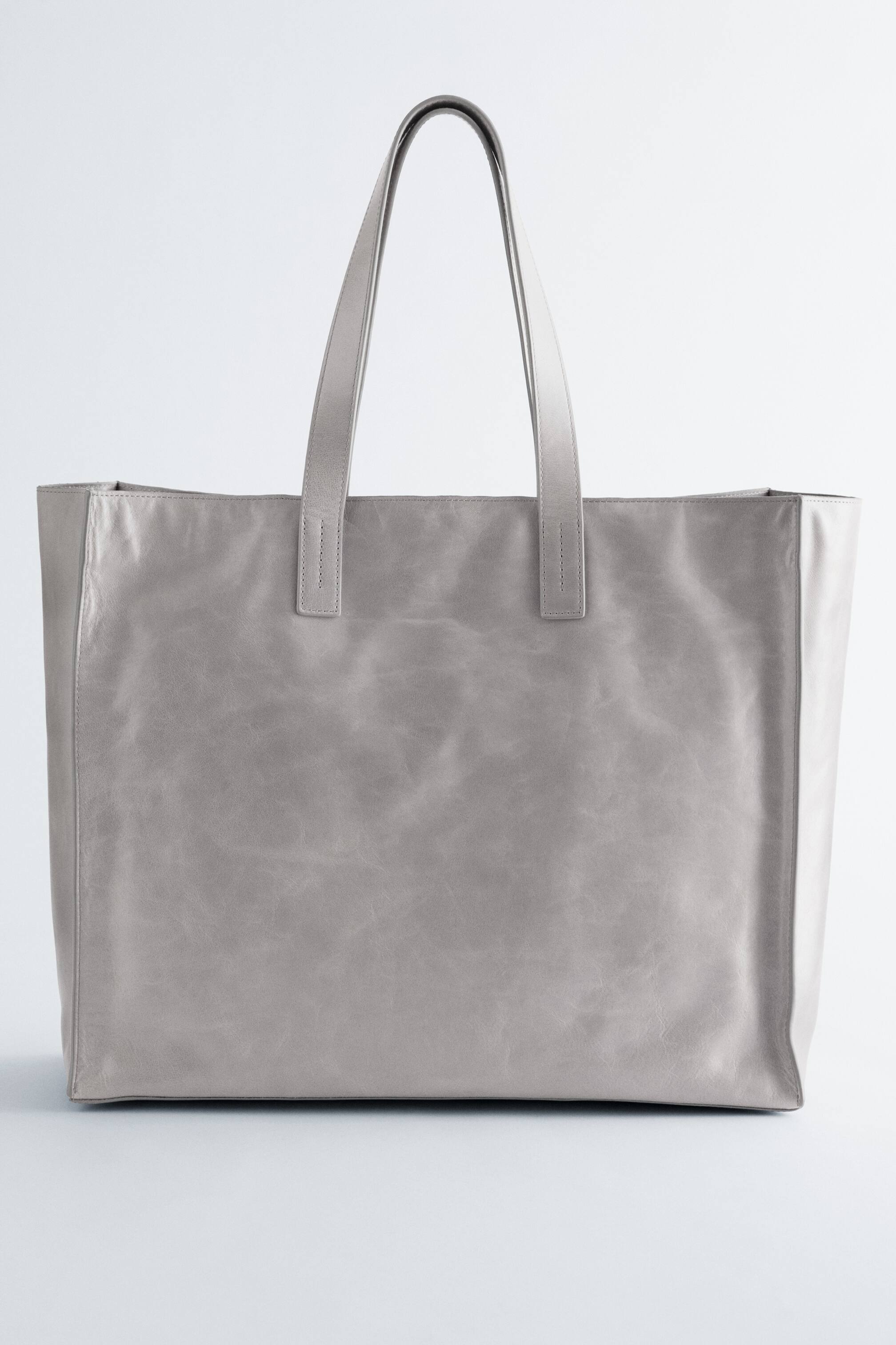 Nagyobb kép megtekintése: Egy világosszürke bőr tote táska látható elölről, amely téglalap alakú és két karcsú felső fogantyúval rendelkezik. A bőr felülete finom textúrát és természetes gyűrődéseket mutat.