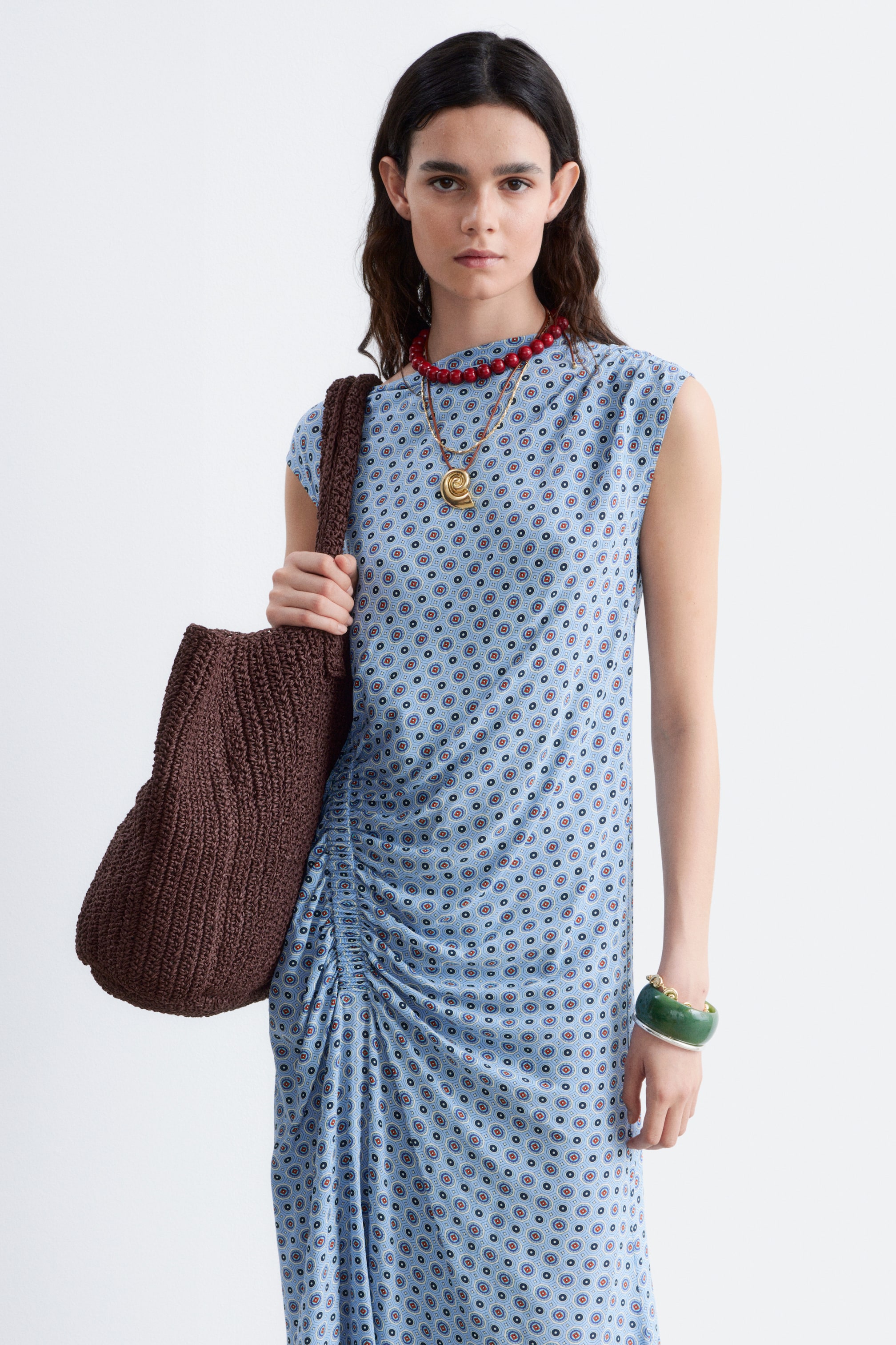 Größeres Bild ansehen: Das hellblaue Kleid besticht durch ein wiederkehrendes Kreismuster, einen hohen Ausschnitt und kurze Ärmel. An der linken Seite ist Raffung eingearbeitet, die Textur schafft und den Midirock hinunterfällt. Eine braune geflochtene Tasche lehnt an der linken Seite.