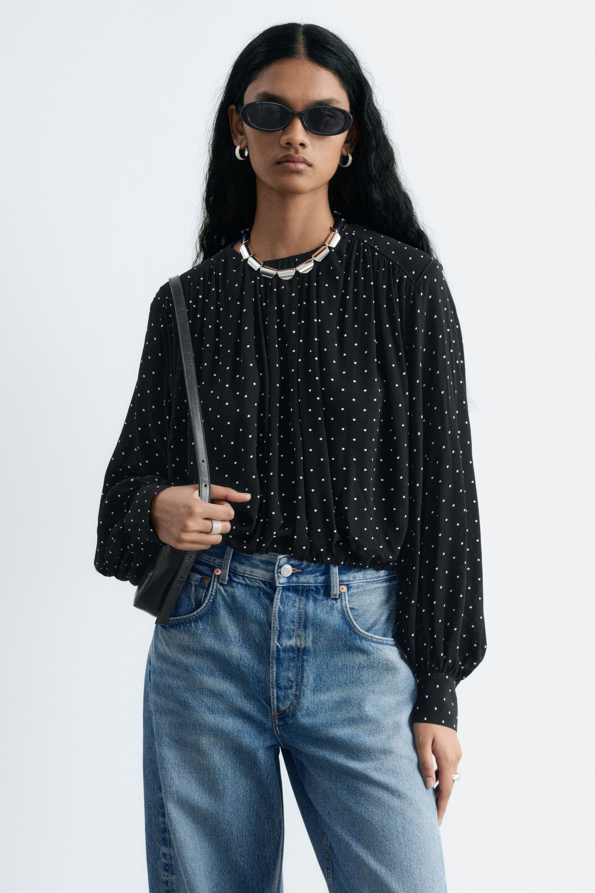 Grotere afbeelding bekijken: Het model draagt een zwarte blouse met een gerimpeld model en een wit polkadotpatroon, ingestopt in een blauwe high-waisted jeans. De blouse heeft een hoge, gerimpelde halslijn en lange, wijdvallende mouwen, en is gestyled met een zwarte schoudertas die het model aan de band vasthoudt.