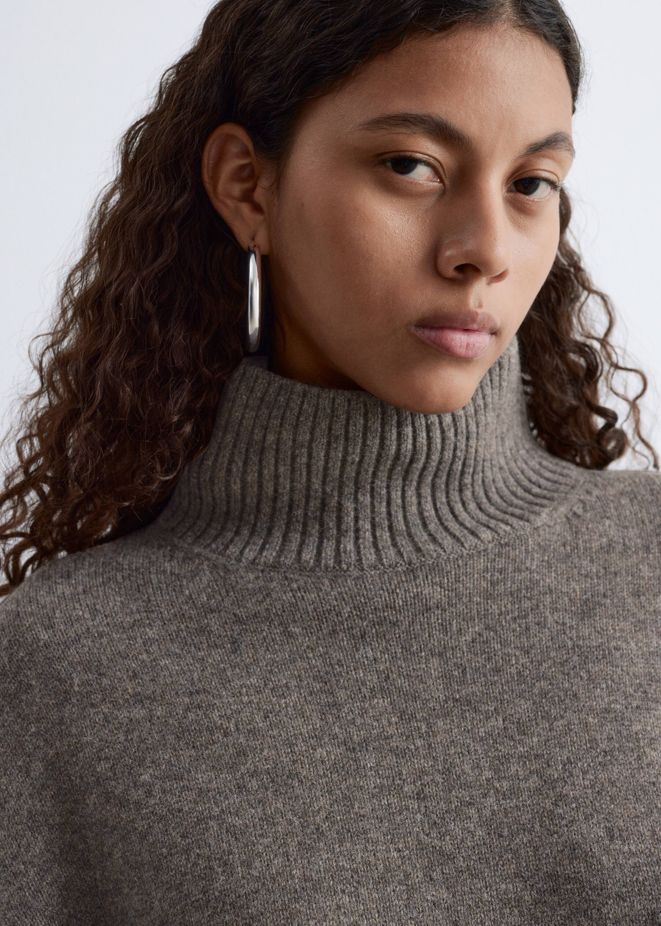 Grösseres Bild ansehen: Das Model trägt einen graubraunen Strickrollkragenpullover mit einem gerippten Kragen und einer weichen, strukturierten Oberfläche.