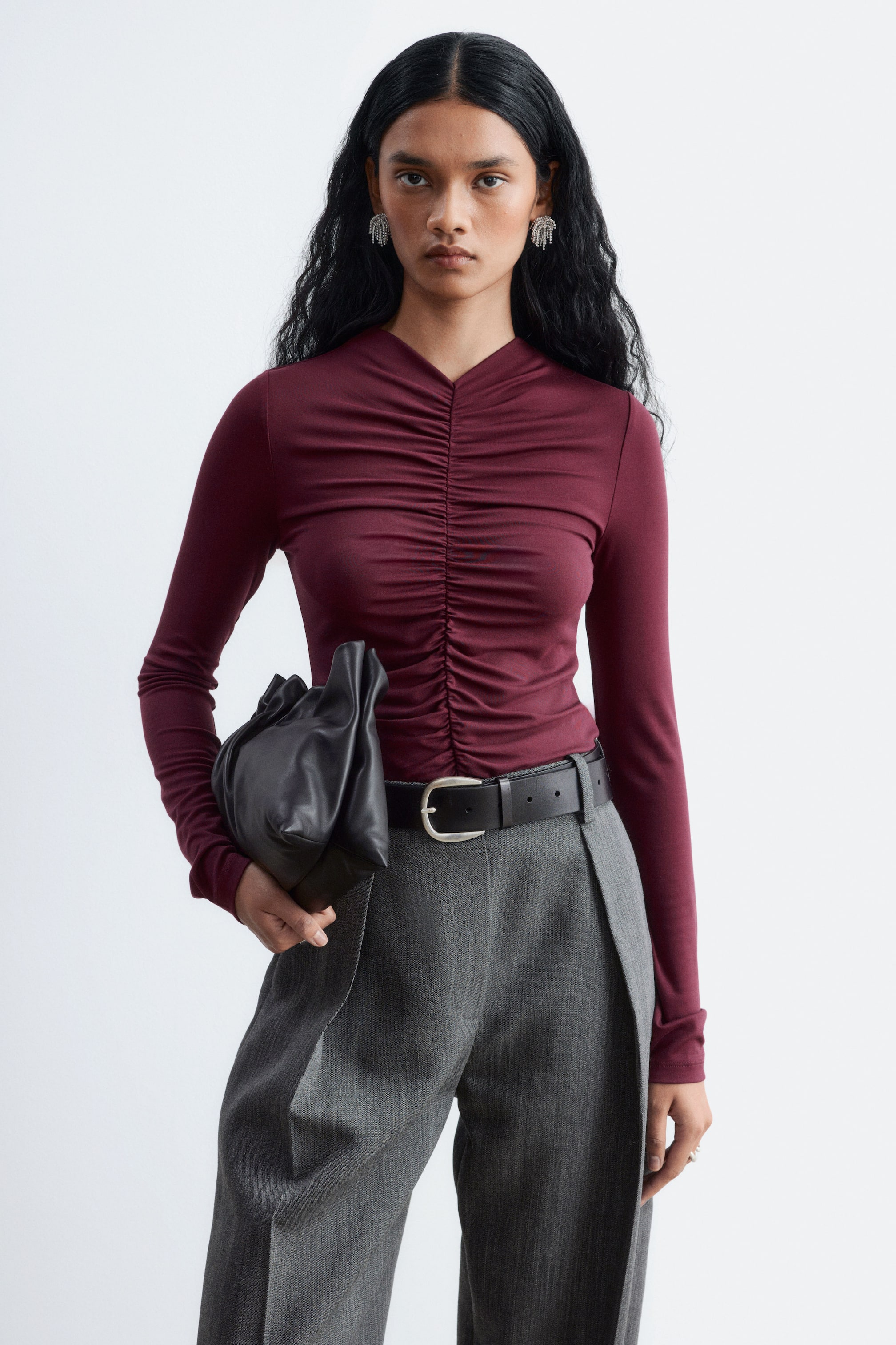 Ver imagem maior: A modelo veste uma camisola de manga comprida cor borgonha, com uma costura franzida no centro da frente, enfiada por dentro de umas calças cinzentas com um cinto de cabedal preto. A modelo está a segurar uma carteira de mão de cabedal preta.
