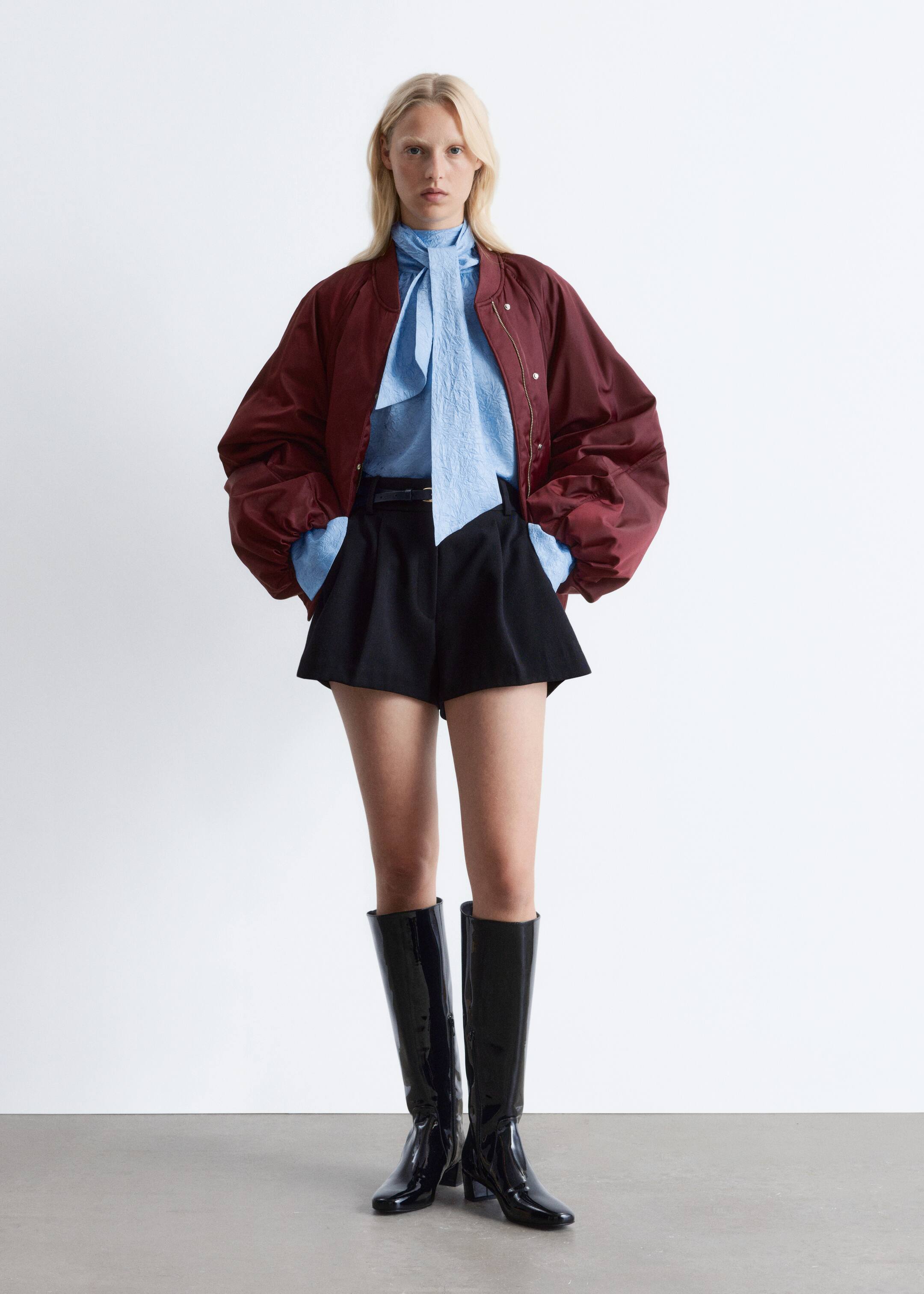 Agrandir l'image: Une veste bomber rouge foncé est portée par-dessus un chemisier à lavallière en tissu froissé bleu clair, glissé dans un short noir à taille haute et évasé, doté d'une ceinture. Le look est complété par des bottes noires en cuir verni montant jusqu'aux genoux, le tout présenté de face.