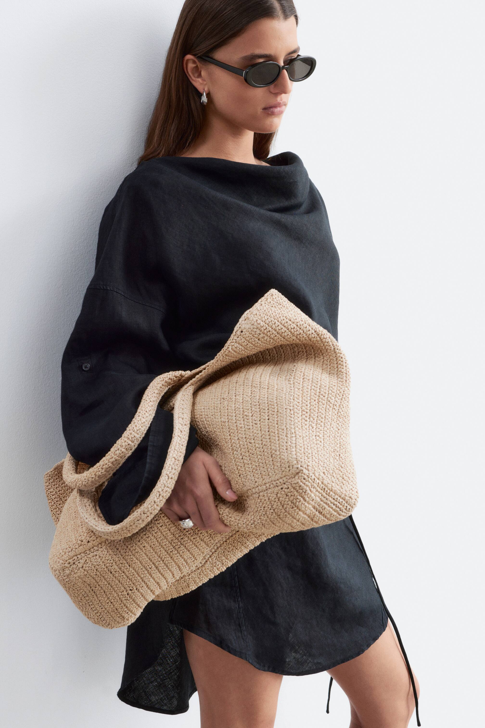 Ver imagem maior: Uma mala de mão em croché bege está a ser segurada por uma mão, exibindo a sua estrutura macia e flexível e a sua textura vertical em croché proeminente. Também apresenta duas alças em croché a condizer, com uma alça a cair sobre a frente da mala.