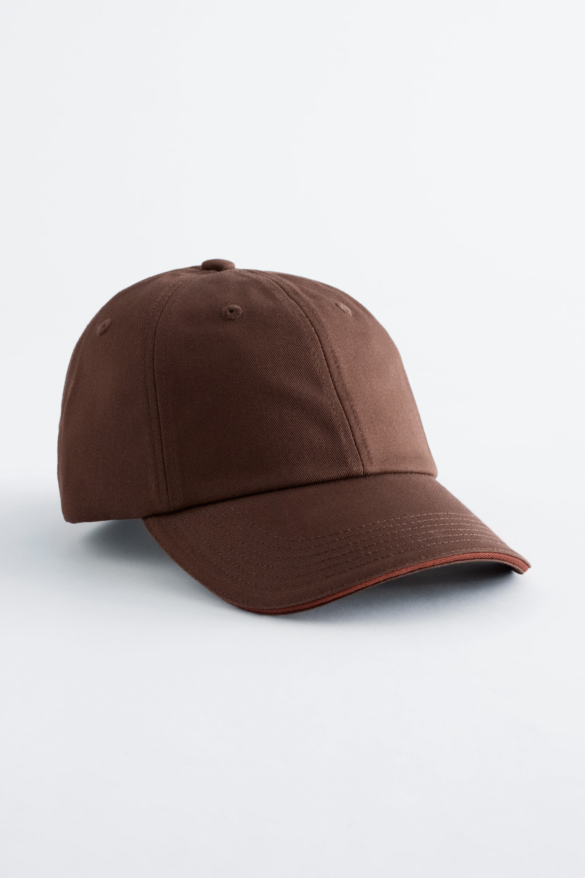 Agrandir l'image: Une casquette de baseball marron, confectionnée en sergé de coton, est inclinée vers l'avant et la droite, affichant un design à panneaux avec des surpiqûres visibles et de petits œillets de ventilation. Elle présente une visière incurvée classique avec des surpiqûres supplémentaires et un passepoil brun-rouge le long de son bord.