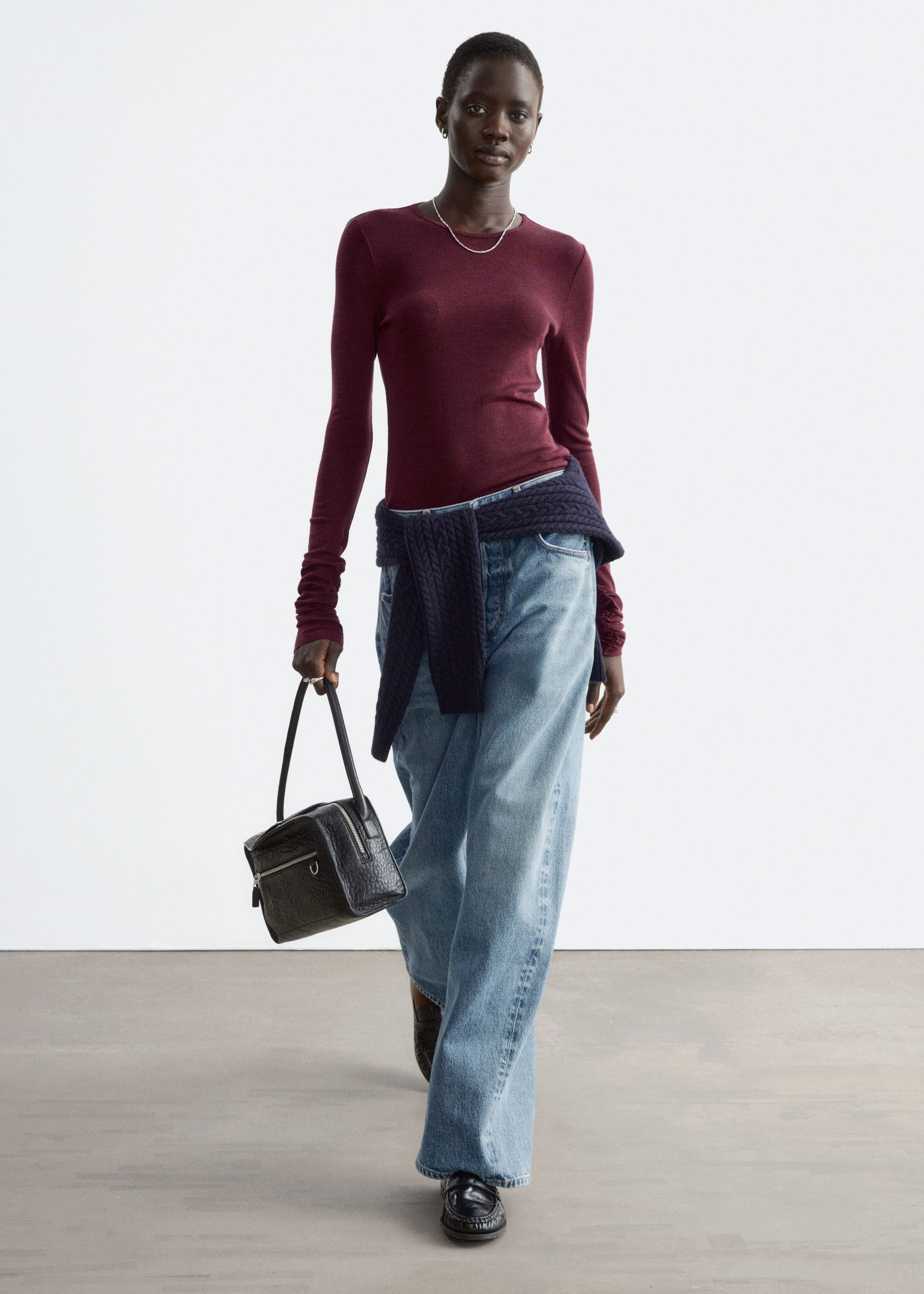 Ein bordeauxrotes, glatt gestricktes Wolloberteil mit Rundhalsausschnitt und langen Ärmeln wird vom Model getragen. Das Oberteil wird mit hellblauen Wide-Leg-Jeans kombiniert, dazu ist ein dunkelblauer Pullover um die Taille gebunden und eine schwarze Lederhandtasche wird getragen.
