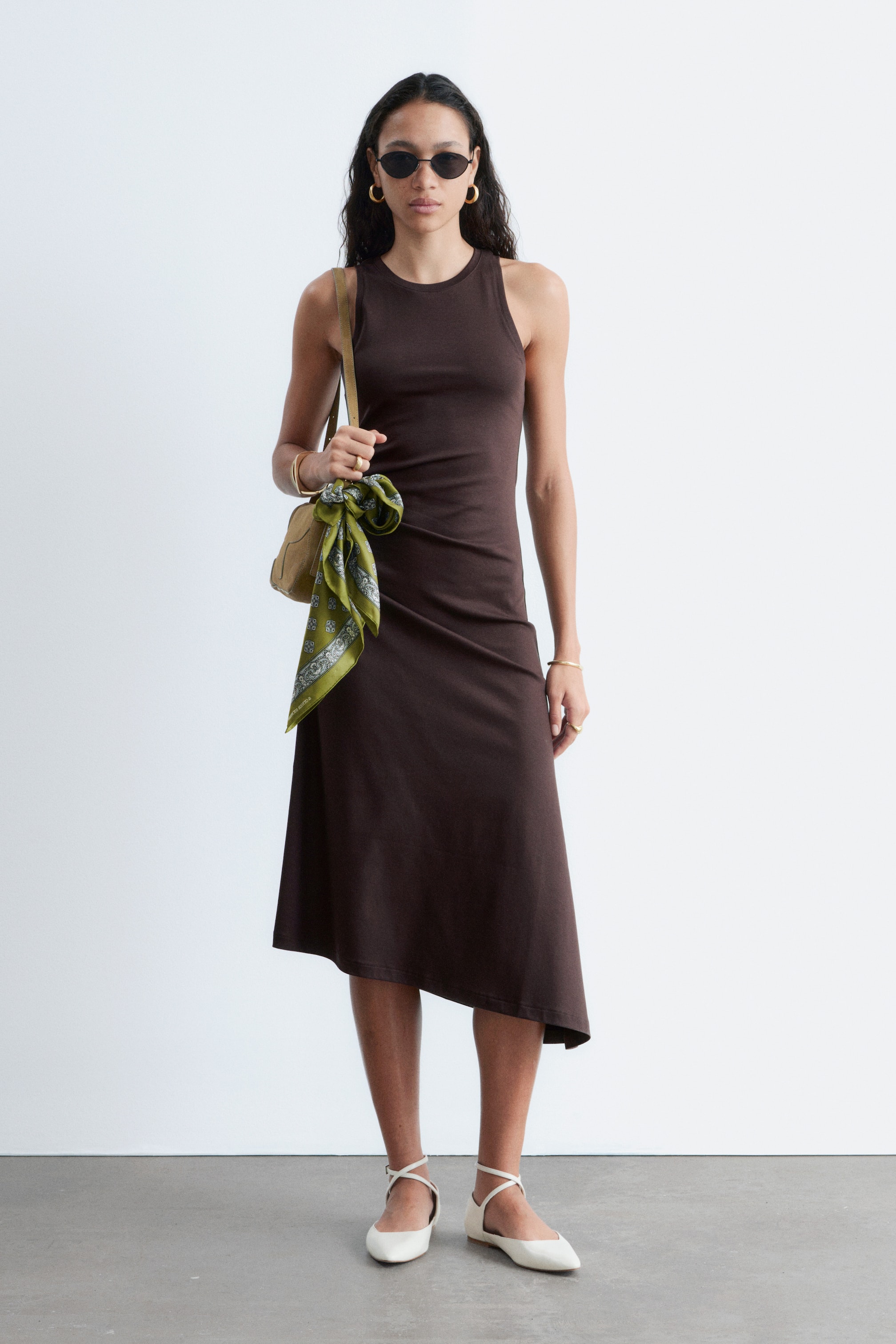 Zobrazit větší obrázek: Tmavě hnědé midi šaty bez rukávů s kulatým výstřihem a přiléhavým živůtkem. Sukně se rozšiřuje do áčkového střihu s asymetrickým lemem, zatímco jemné řasení zdobí levou stranu pasu.