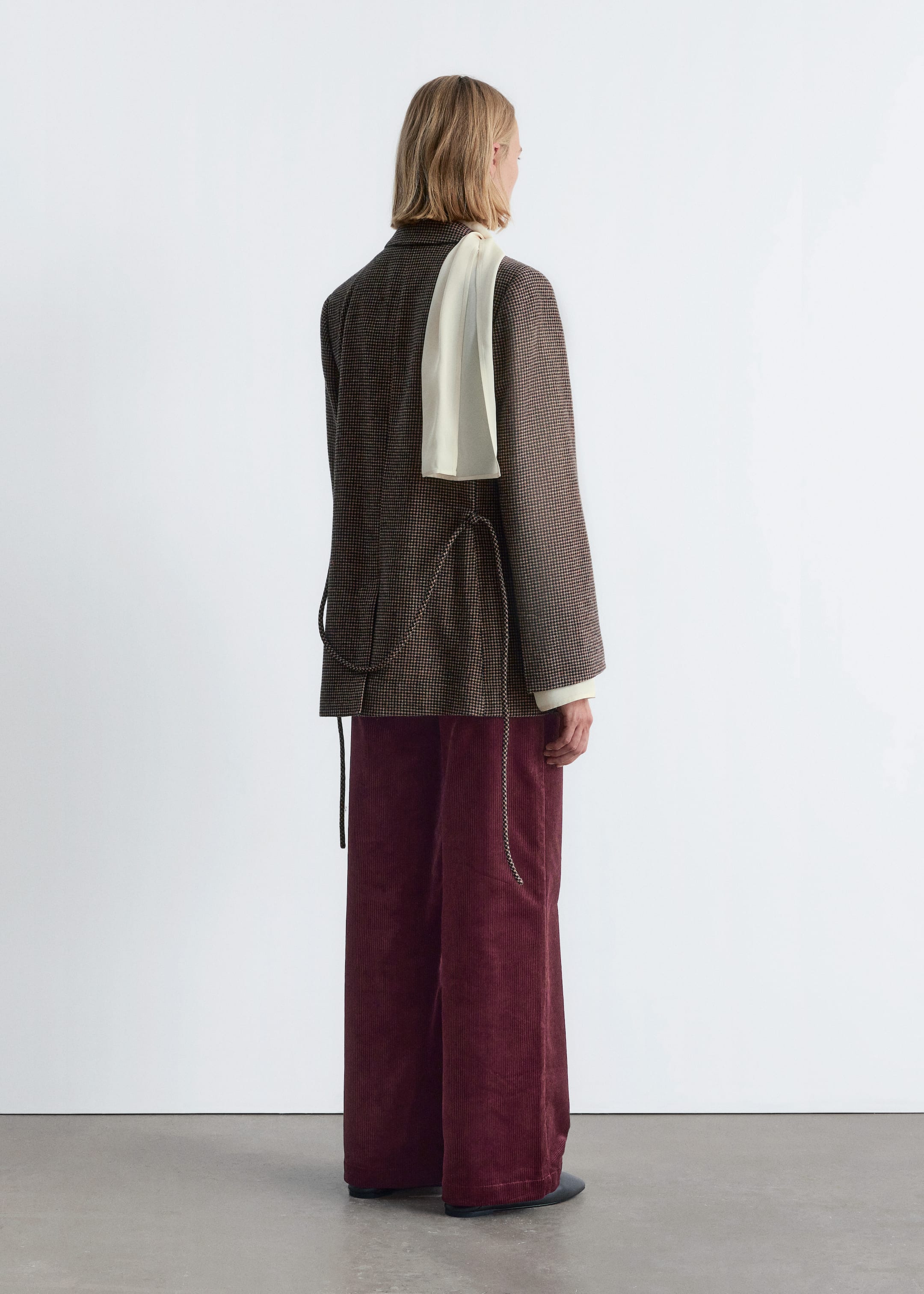 Größeres Bild anzeigen: Eine braun-schwarze Hahnentrittjacke wird von hinten und von der rechten Seite präsentiert, mit einem Kordelzug, der durch den unteren Rücken gefädelt ist und herabhängende Enden hat. Ein weißes Langarmshirt wird darunter getragen, kombiniert mit locker sitzenden, weiten burgunderroten Cordhosen. Ein cremeweißer Schal wird über die linke Schulter drapiert.