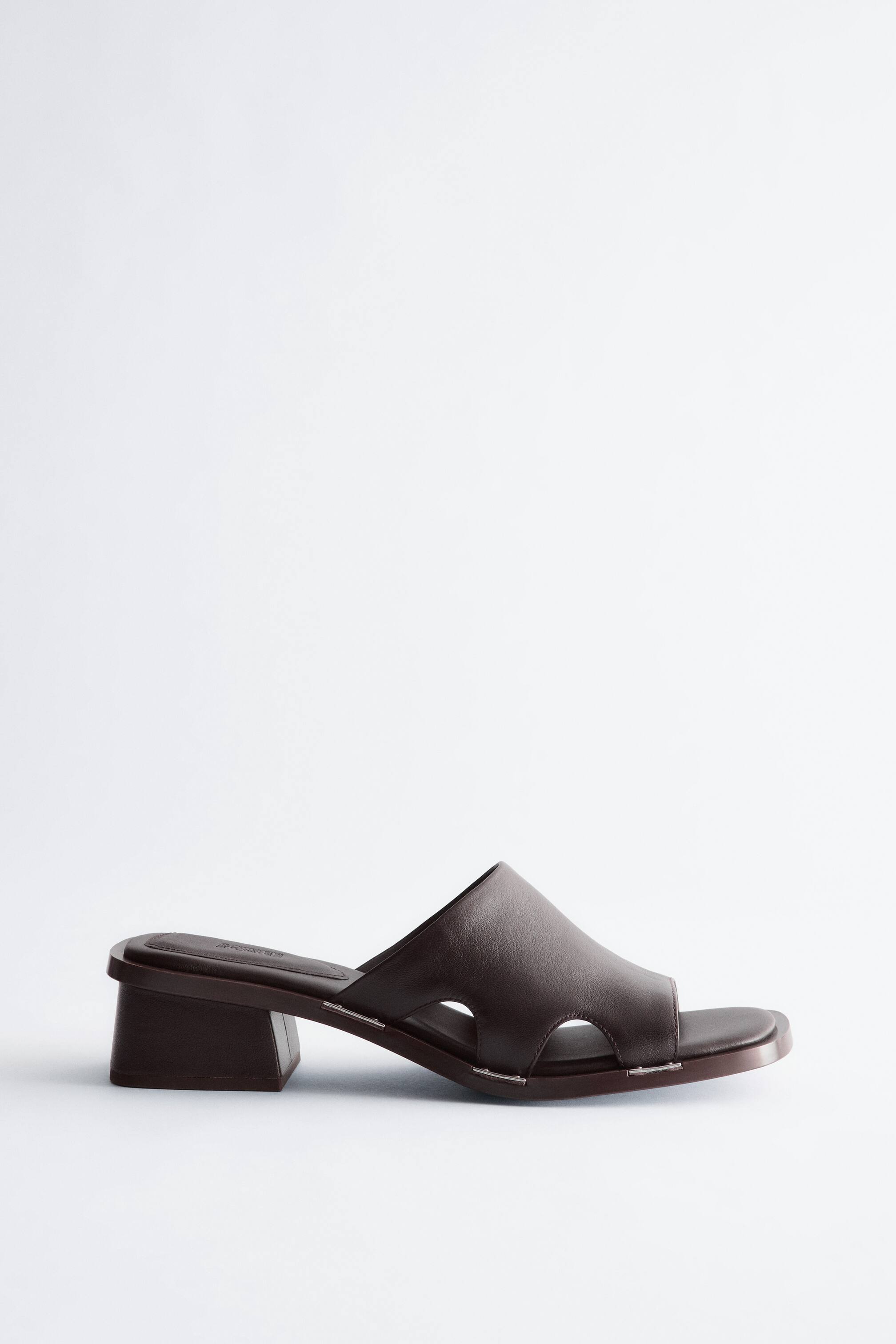 Προβολή μεγαλύτερης εικόνας: Ένα σκούρο καφέ δερμάτινο mule σανδάλι είναι στραμμένο προς τα αριστερά, διαθέτοντας ένα φαρδύ λουράκι στο μπροστινό μέρος με δύο πλευρικά κοψίματα. Δείχνει ένα τετράγωνο ανοιχτό δάχτυλο και ένα χαμηλό τετράγωνο τακούνι, με μικρά ασημί συνδετήρες που ασφαλίζουν το λουράκι στη σόλα.