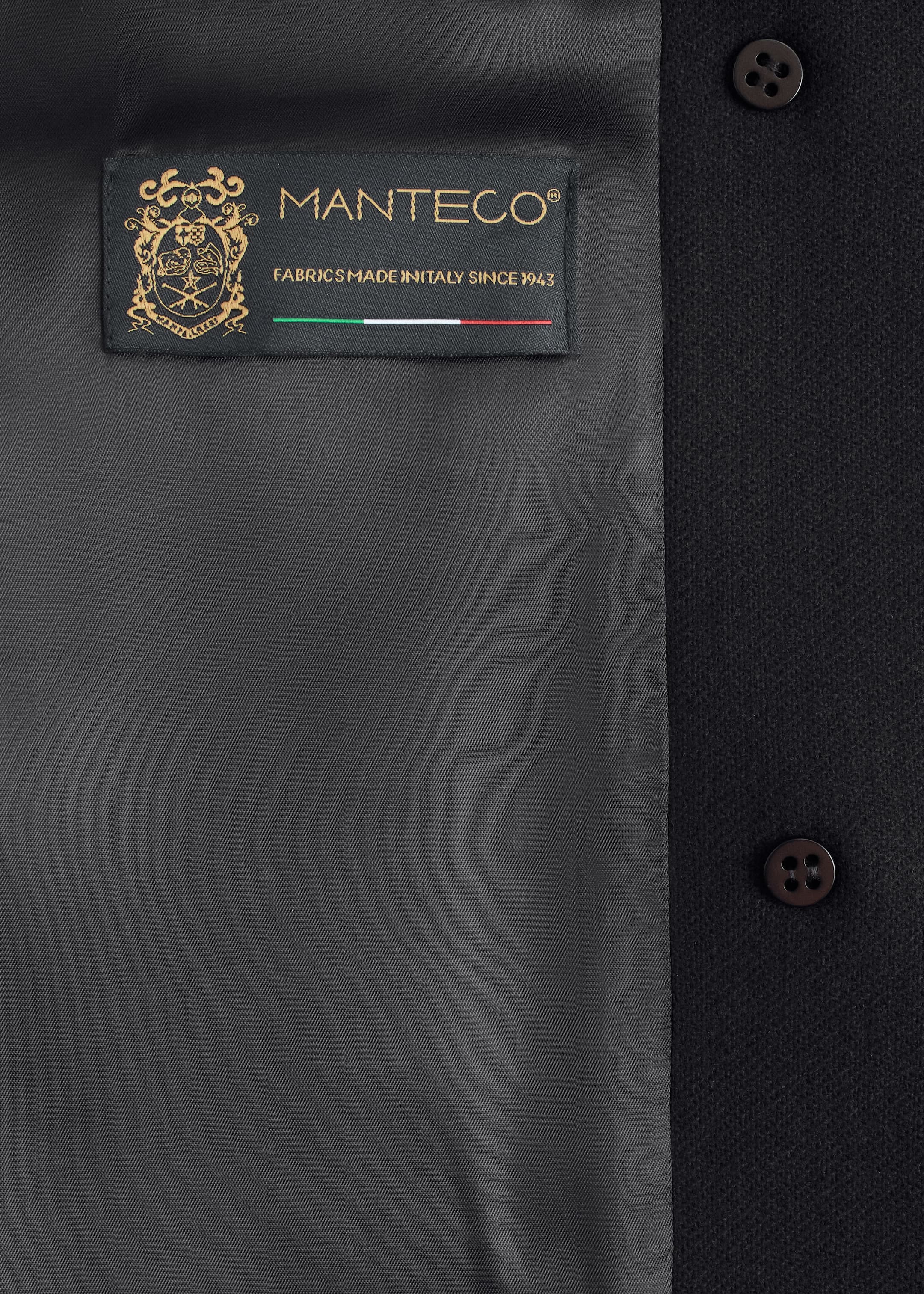 View larger image: A black wool-blend coat is being shown in a close-up, revealing its dark gray lining on the left and the texture of its outer fabric on the right. A black rectangular label with gold 'MANTECO' text and an Italian flag is being stitched onto the lining, and two dark brown, four-hole buttons are visible on the outer fabric.