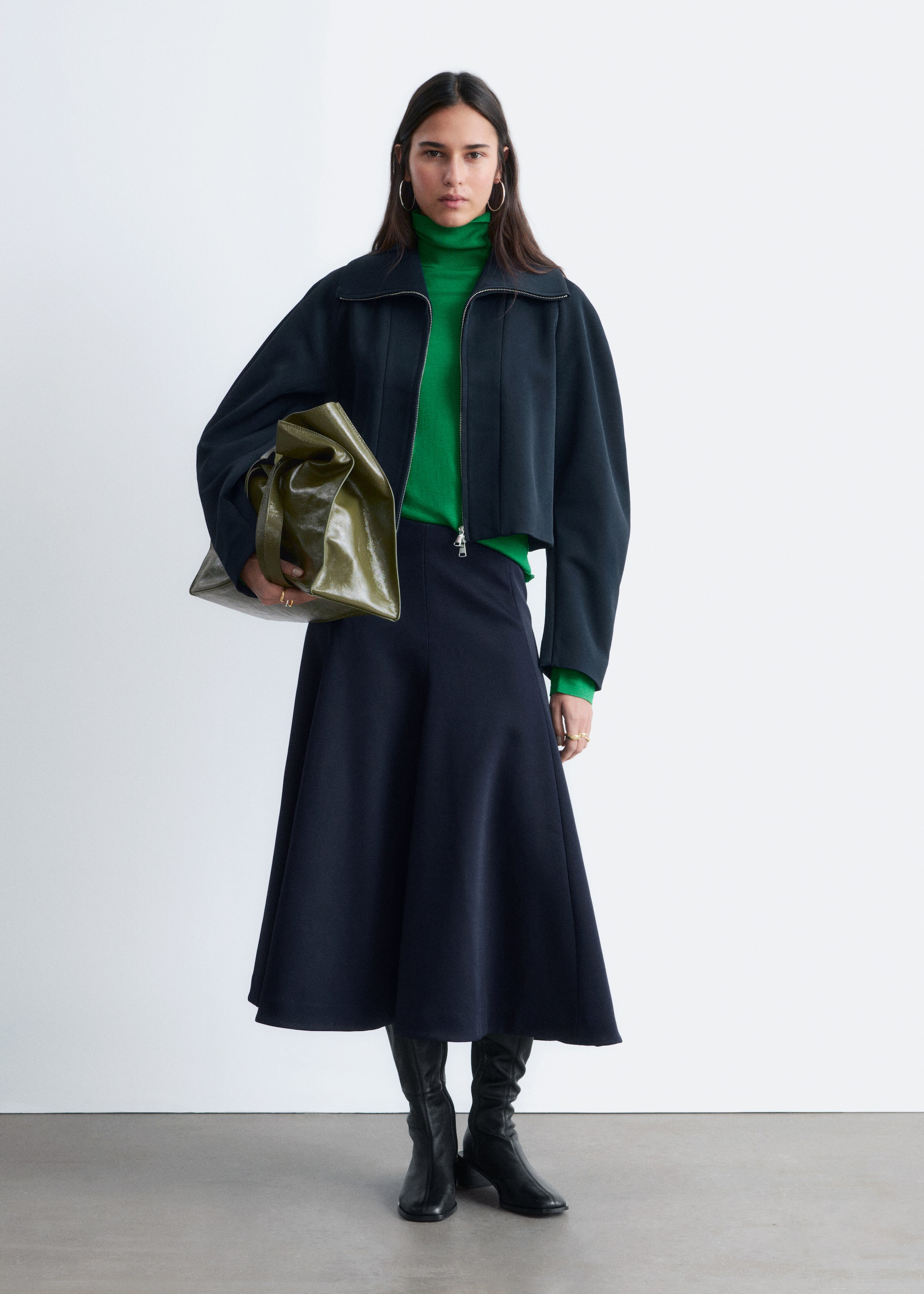 View larger image: A dark blue cropped jacket features a silver front zip and a wide collar, layered over a bright green polo neck. A matching dark blue flared midi skirt extends to mid-calf, complementing black knee-high boots, while a shiny olive green unstructured handbag is carried.