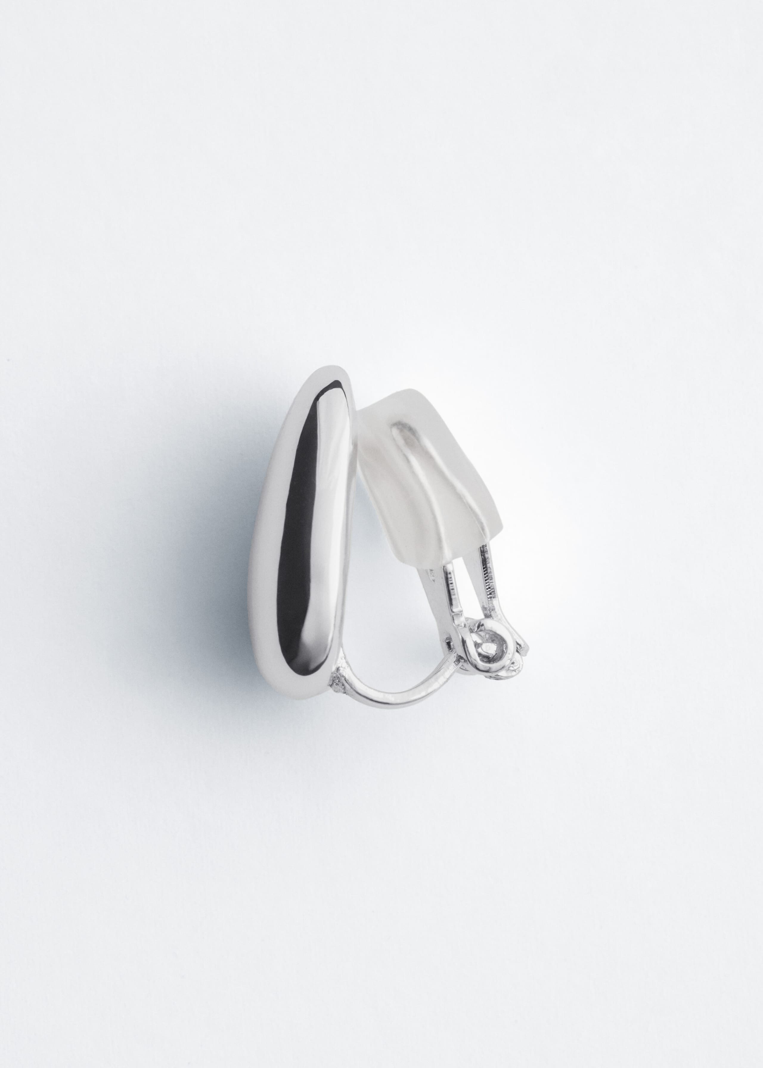 A silver-toned clip-on earring featuring a polished, sculptural teardrop-shaped front is shown in side profile, displaying its hinge mechanism and a translucent silicone comfort pad.