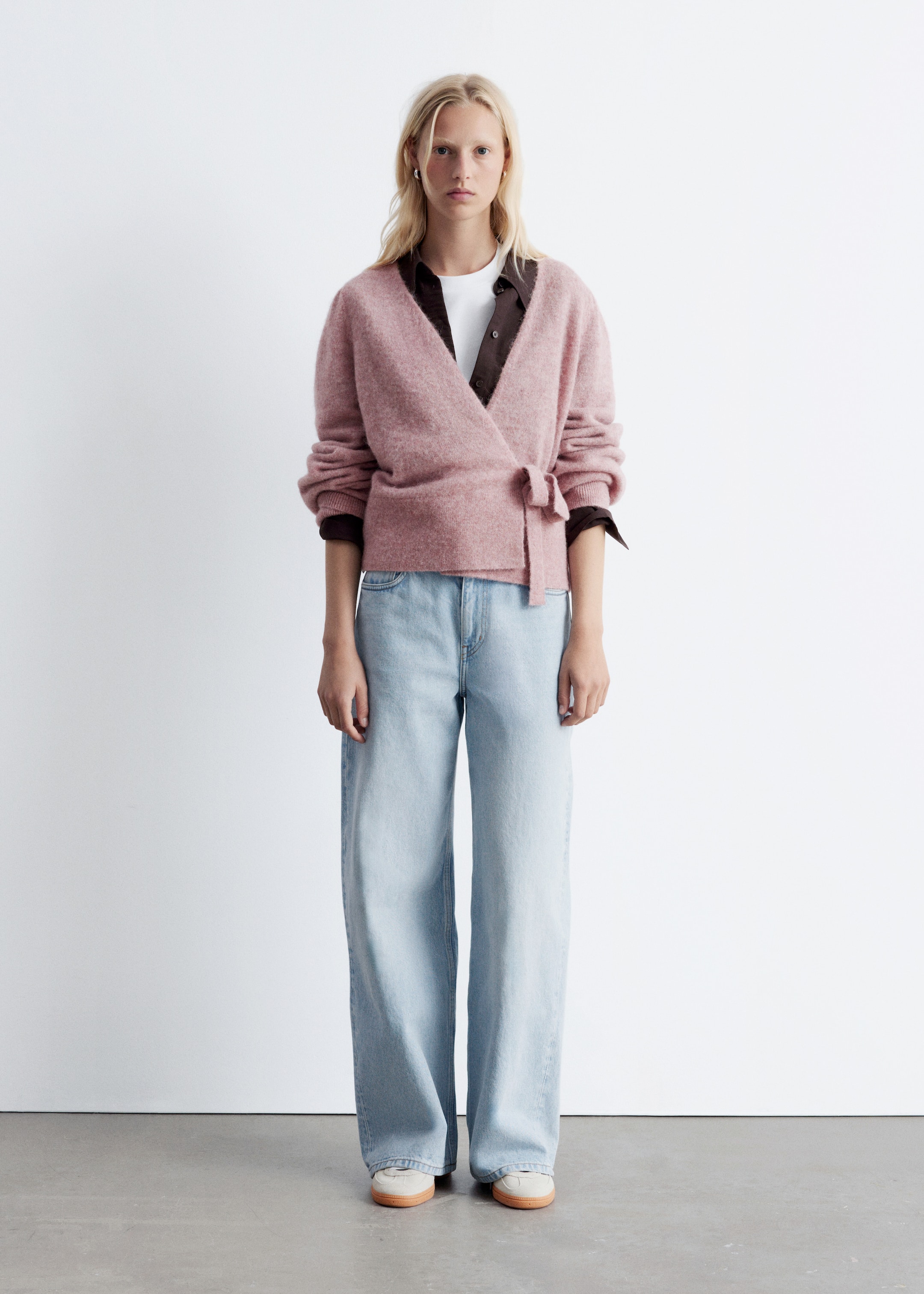 Ver imagem maior: A modelo está a usar um casaco de malha rosa com decote cruzado, mangas compridas bufantes, punhos e bainha canelados, e um fecho de amarrar na lateral, sobreposto a uma t-shirt branca e uma camisa escura com colarinho.