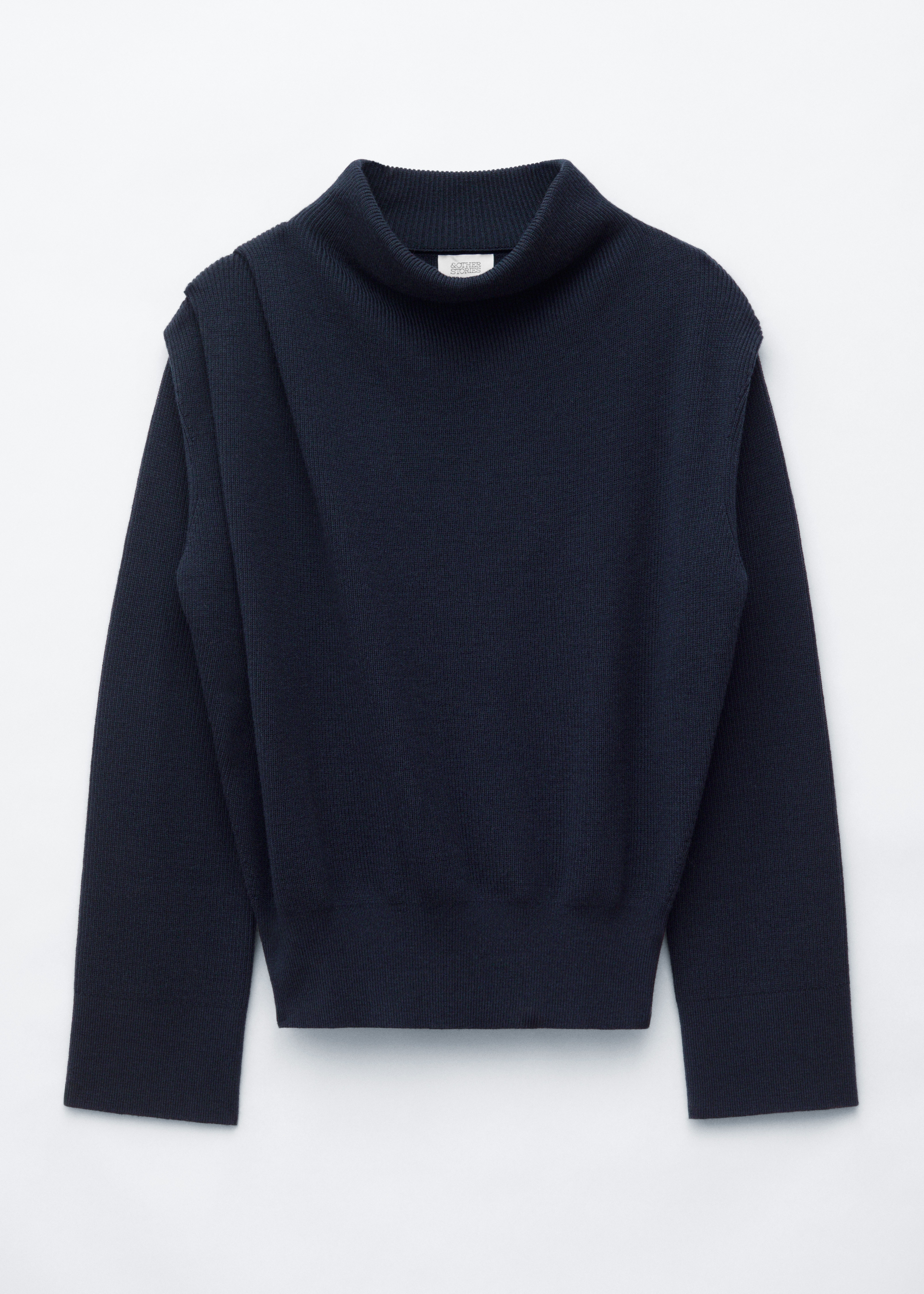 Un maglione in lana a coste blu navy con scollo a cowl alto e drappeggiato e maniche lunghe Ã¨ indossato dalla modella. Il maglione Ã¨ abbinato a jeans in denim scuro, mentre la modella porta una borsa a tracolla in pelle marrone scuro su un braccio.