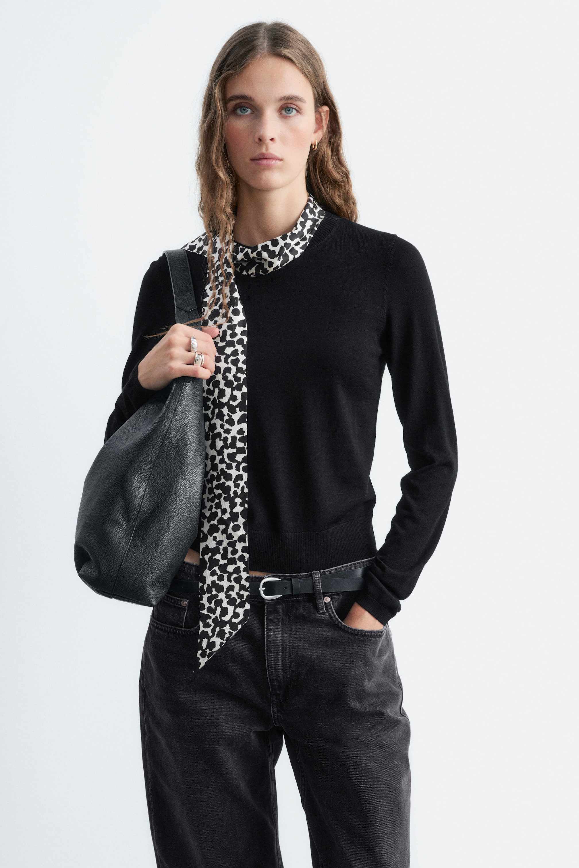 Nagyobb kép megtekintése: A Silk Collar Wool Jumper modellt viseli, amely egy fekete, finom kötésű, kerek nyakú fazont mutat, elöl lazán leomló, integrált fekete-fehér mintás selyemsállal. A pulóvert sötét farmerrel, fekete övvel és az egyik vállon viselt fekete bőr válltáskával stylingolták.