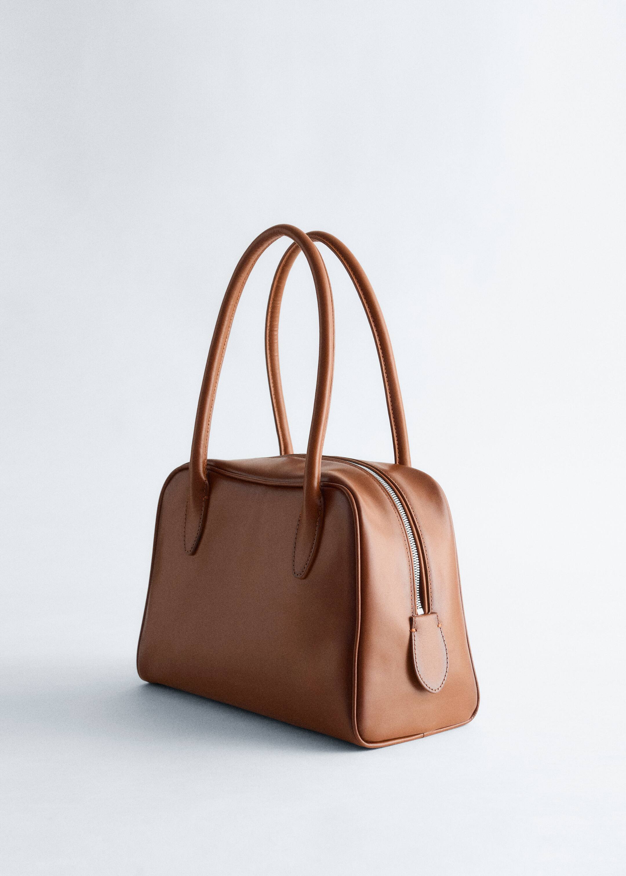 Größeres Bild ansehen: Eine braune Bowlingtasche aus glattem Leder steht da und präsentiert ihren strukturierten Körper, zwei abgerundete obere Griffe und einen silberfarbenen Reißverschluss mit einem passenden Zugband entlang der Oberseite.