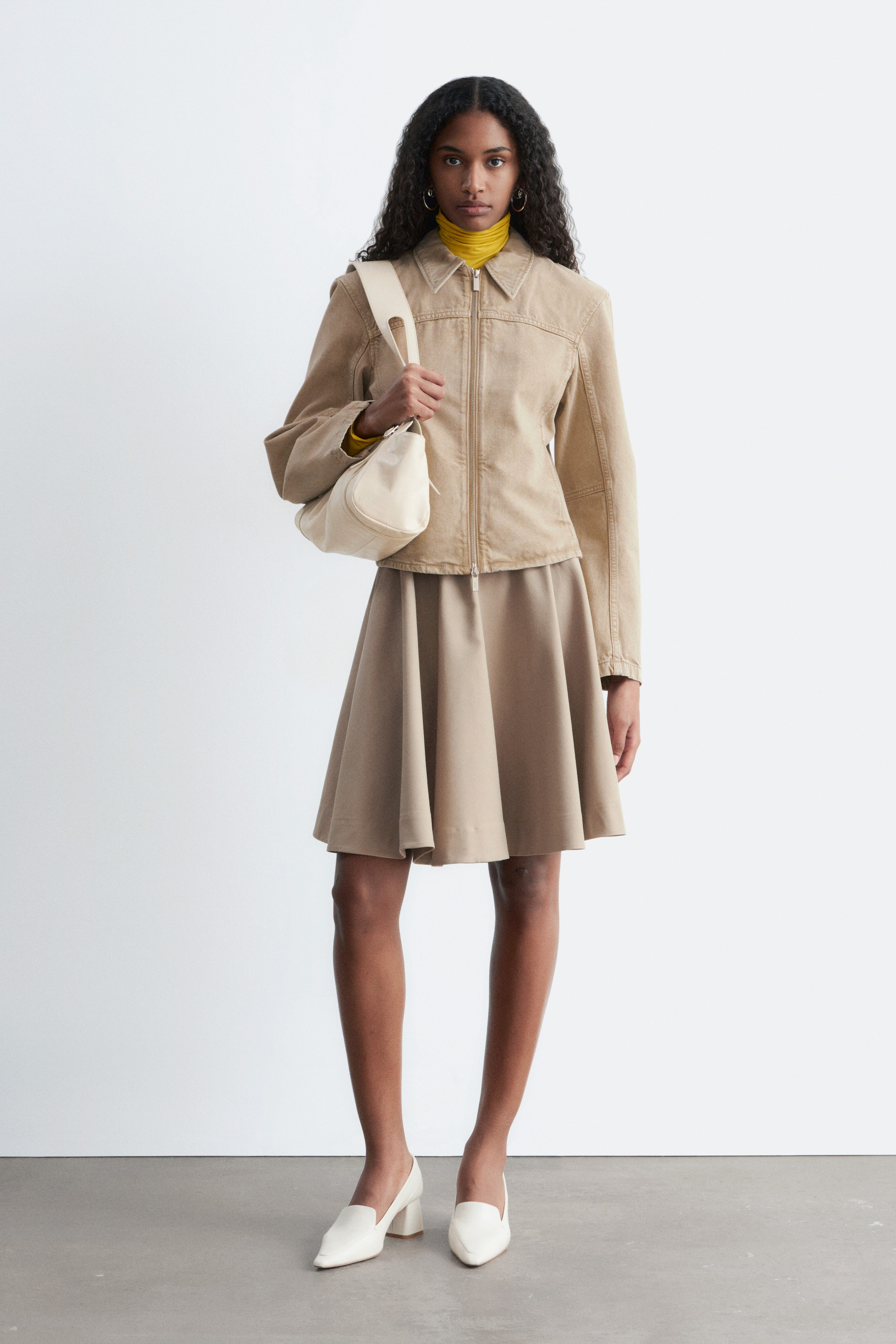 Zobraziť väčší obrázok: Modelka má na sebe hnedú džínsovú bundu, ktorá sa vyznačuje priliehavým strihom, tvarovanými rukávmi a predným zapínaním na zips. Jemné vrásnenie je viditeľné na švoch a bunda je vrstvená na žltom roláku.