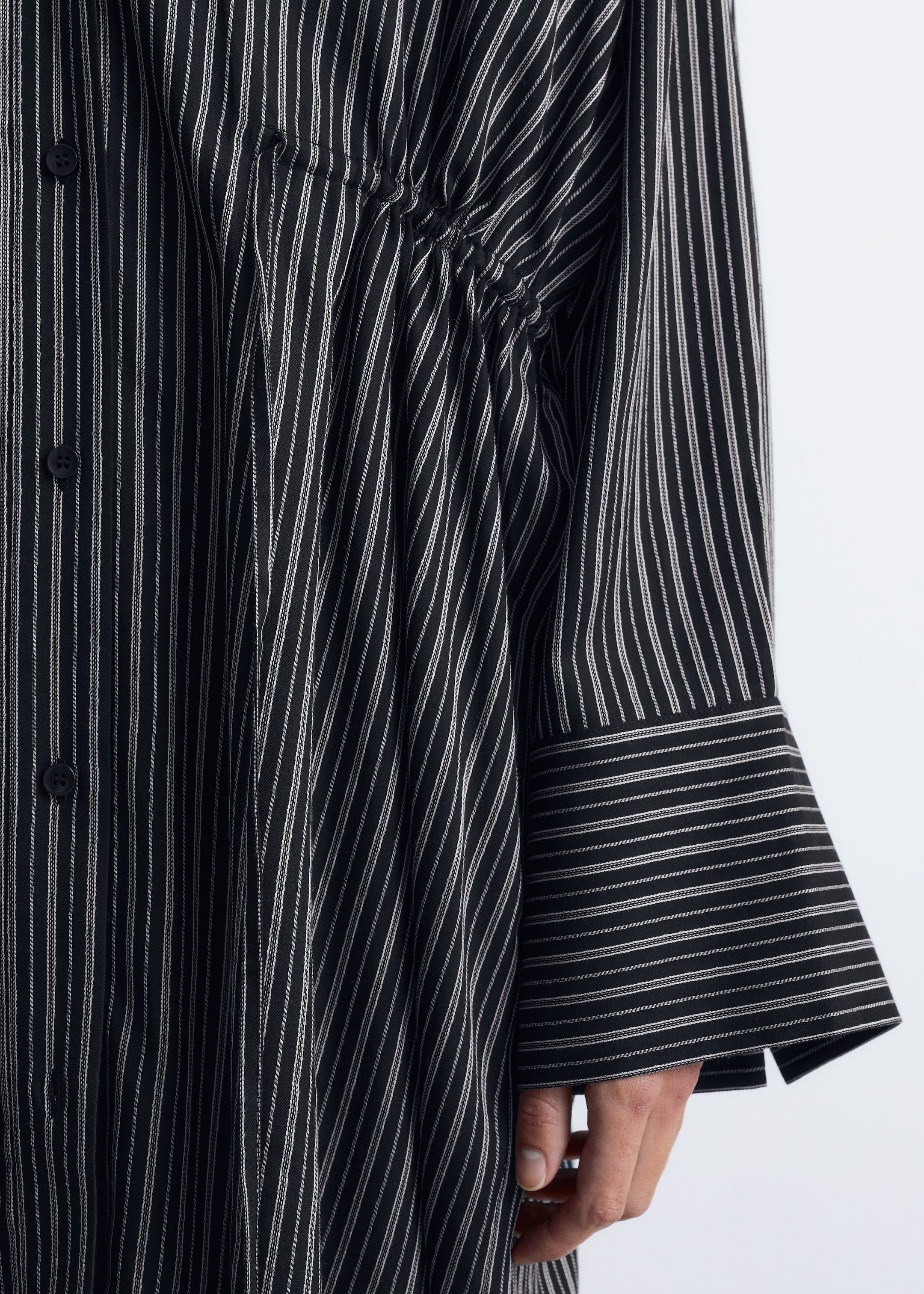 Größeres Bild anzeigen: Ein schwarzes Hemdkleid mit dünnen weißen vertikalen Streifen wird von dem Model getragen, es verfügt über eine geraffte Kordelzug-Taille, die Falten erzeugt, sowie lange Ärmel und breite Manschetten.
