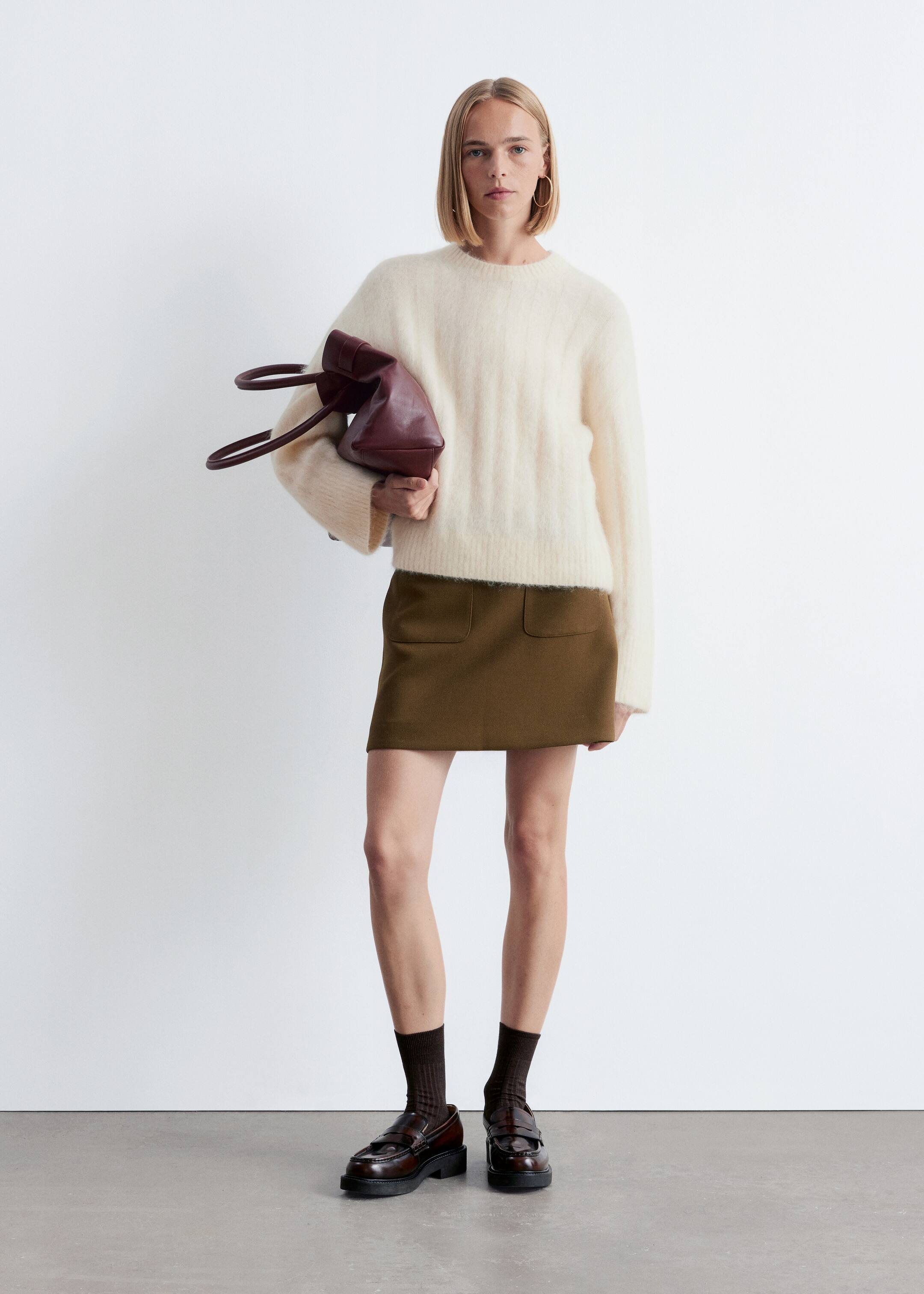Se større billede: Et outfit bliver præsenteret, med en cremefarvet, fuzzy crew-neck sweater båret over en olivengrøn mini nederdel med to forlommer. Mørkebrune ribbede sokker styles med mørkebrune chunky-sålede loafers, og en mørkerød struktureret taske bliver holdt.
