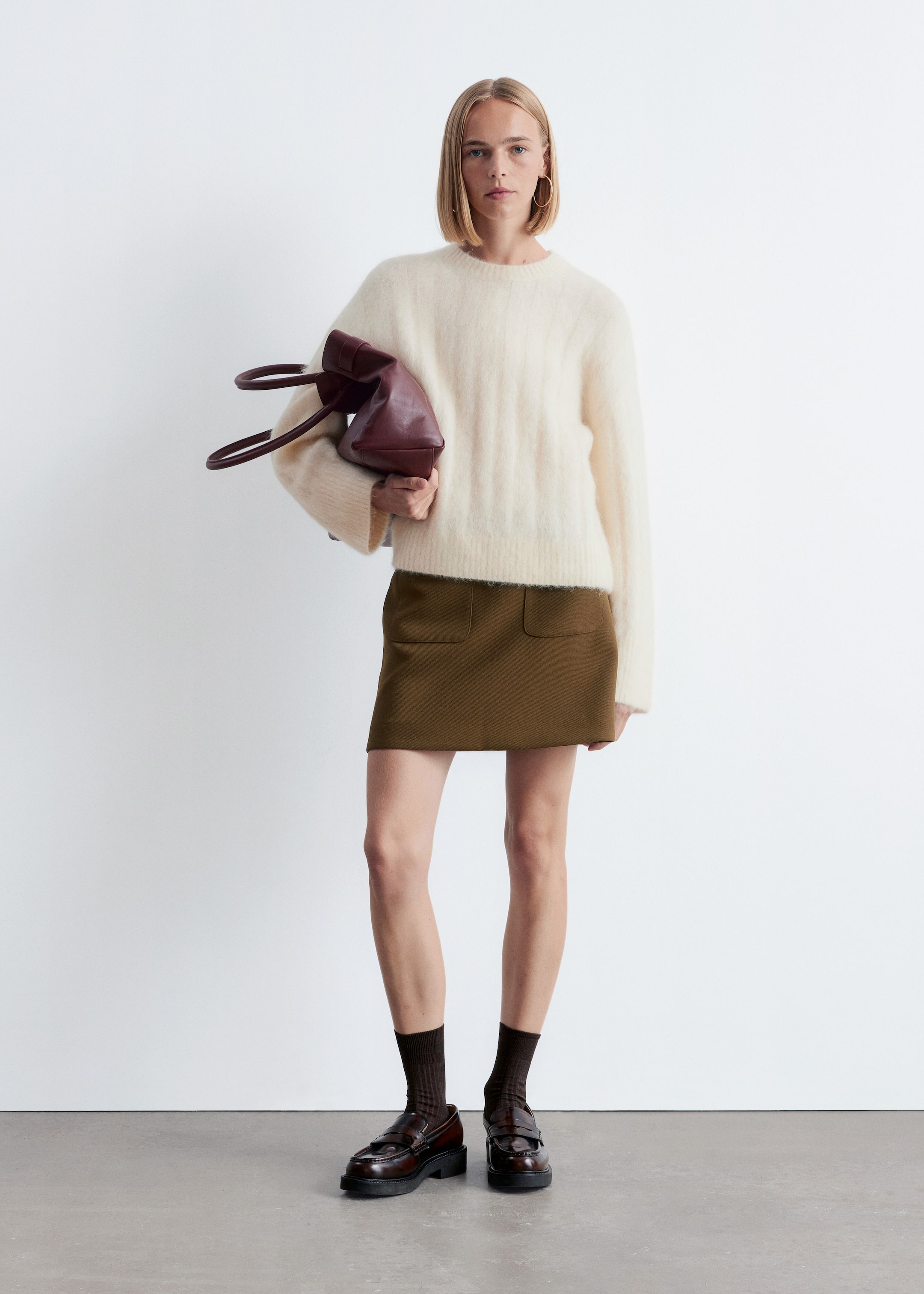 View larger image: An outfit is being presented, featuring a cream-colored, fuzzy crew-neck sweater worn over an olive mini skirt with two front patch pockets. Dark brown ribbed socks are being styled with dark brown chunky-soled loafers, and a dark red structured bag is being held.