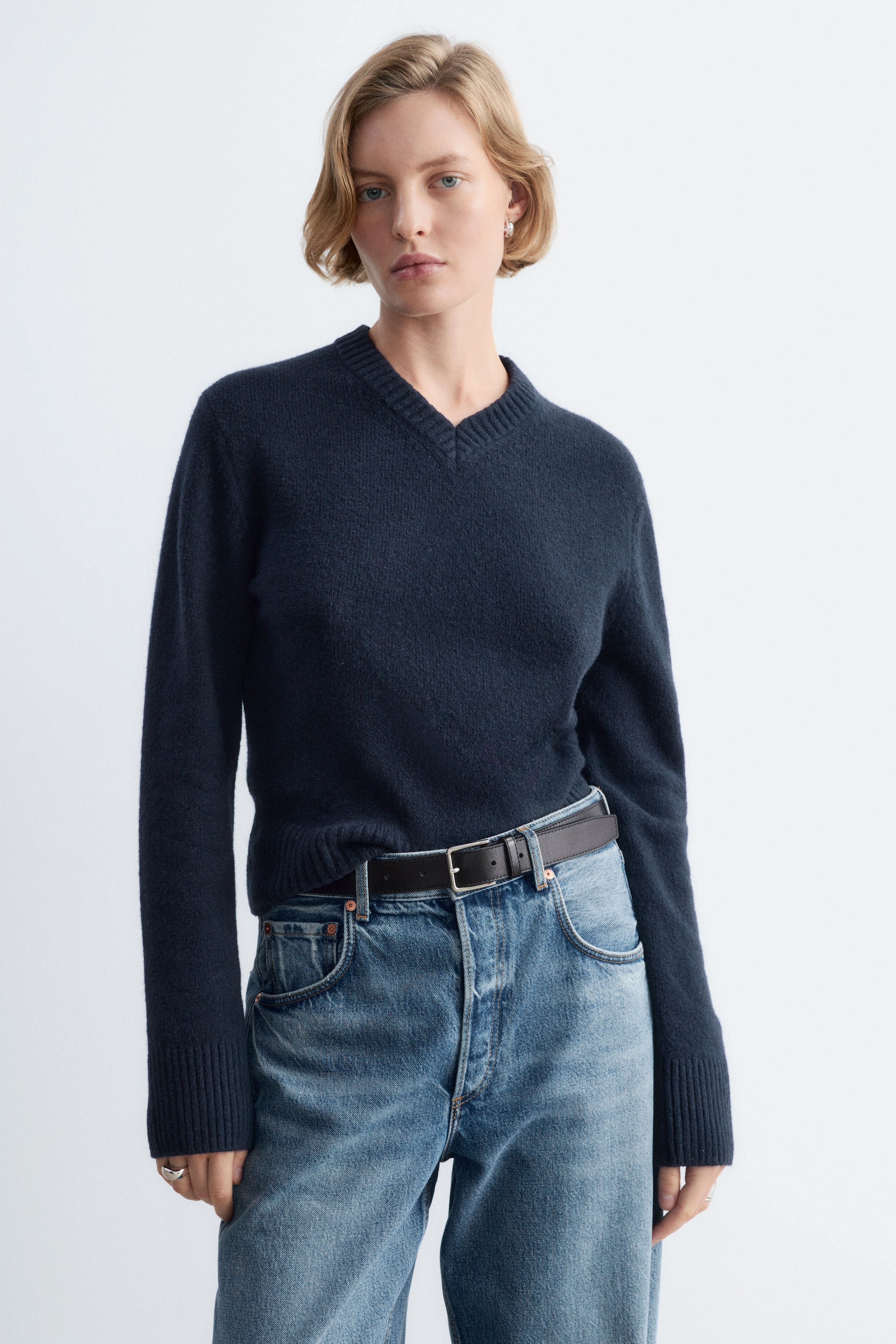 Visa större bild: En marinblå stickad tröja med ribbad V-ringning och ribbade manschetter bärs av modellen. Tröjan är instoppad i ett par blå jeans som hålls på plats med ett svart bälte.