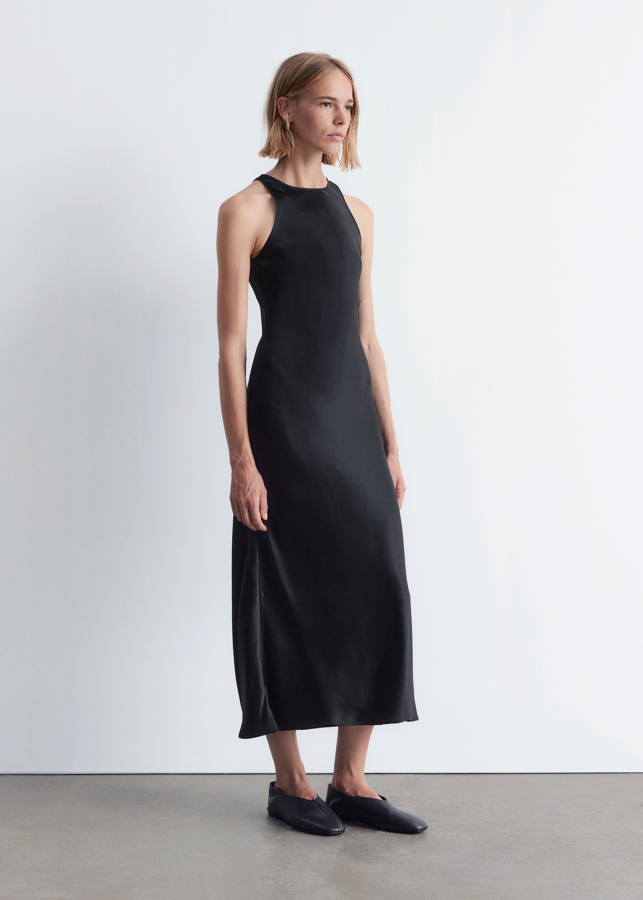 Grösseres Bild ansehen: Ein schwarzes Midikleid wird von einem Model getragen, das nach rechts schaut und den hohen runden Ausschnitt, die eingeschnittenen Armlöcher und die fließende, säulenartige Silhouette zeigt, die bis zur Mitte der Wade reicht.