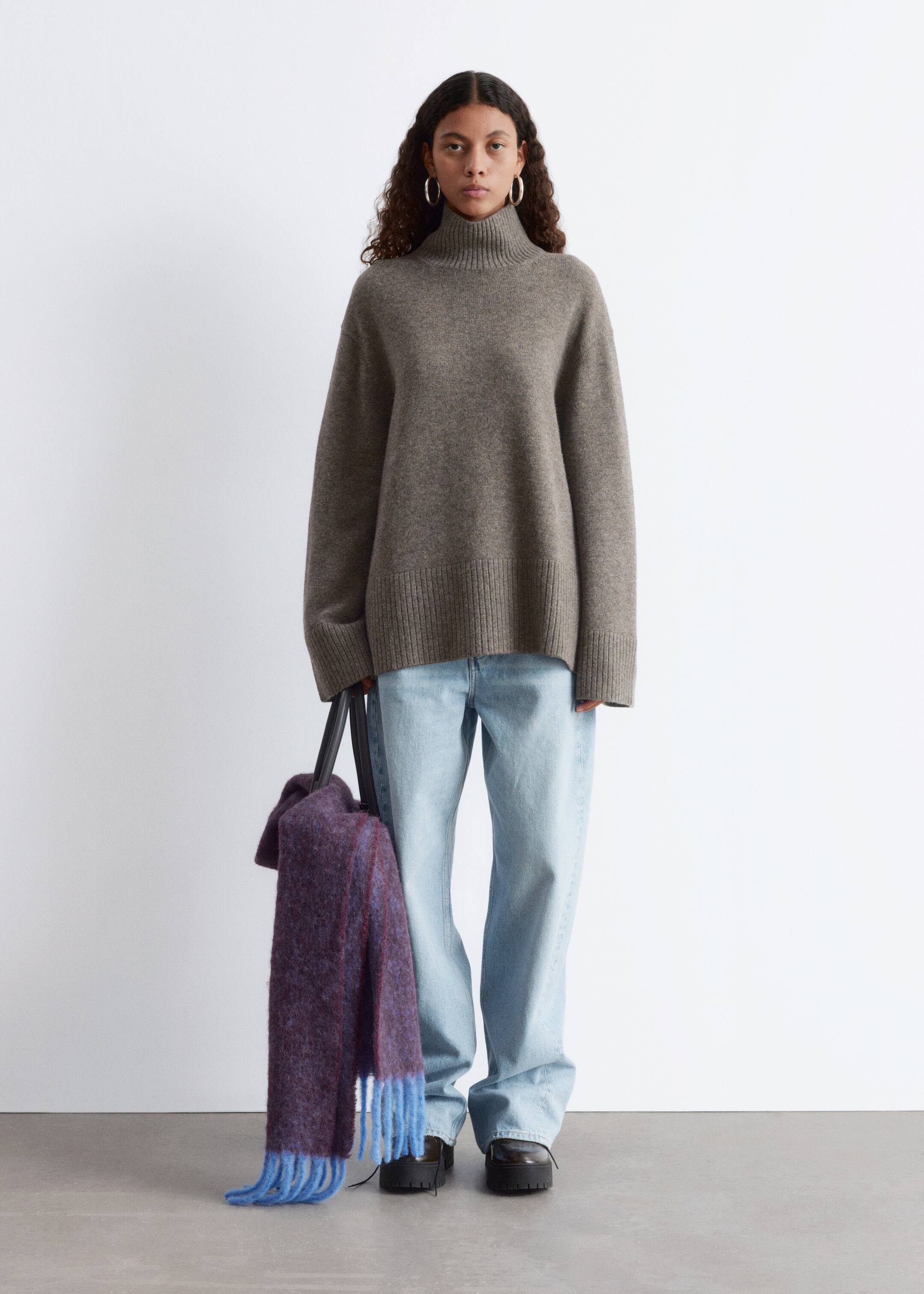 Grösseres Bild ansehen: Ein grauer, locker sitzender Rollkragenpullover mit geripptem Kragen und geripptem Saum wird nach vorne gerichtet präsentiert und mit einer hell gewaschenen, weit geschnittenen Jeans und schwarzen Schuhen mit klobigen Sohlen kombiniert. Ein strukturierter lila Schal mit blauen Fransen hängt von einer schwarzen Tasche und vervollständigt das Ensemble.