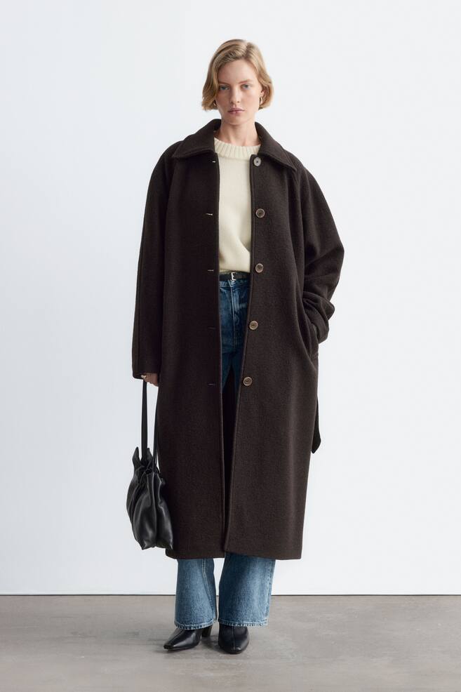 Ein langer, dunkelbrauner Wollmantel mit weitem Kragen und Knopfleiste wird vom Model offen getragen. Der Mantel ist Ã¼ber einem cremefarbenen Rundhalspullover und einer blauen Jeans kombiniert, wÃ¤hrend in einer Hand eine schwarze Lederhandtasche gehalten wird. - 1