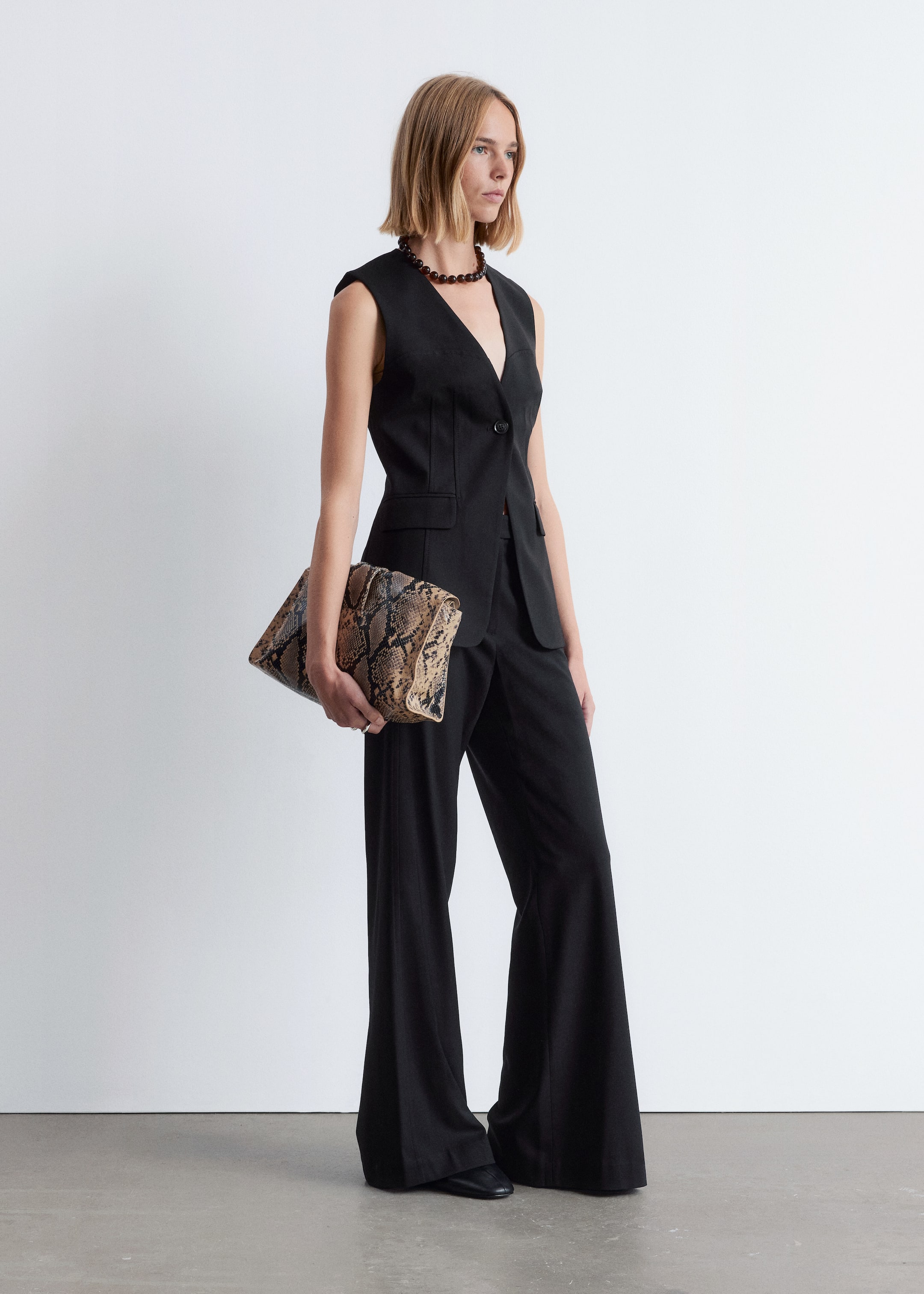 Agrandir l'image: Un gilet noir sans manches et un pantalon large assorti sont présentés comme un ensemble coordonné, le gilet présentant un décolleté en V profond, une fermeture à un bouton et deux poches à rabat. Une pochette à imprimé serpent est tenue, et un collier ras du cou en perles sombres orne le cou.