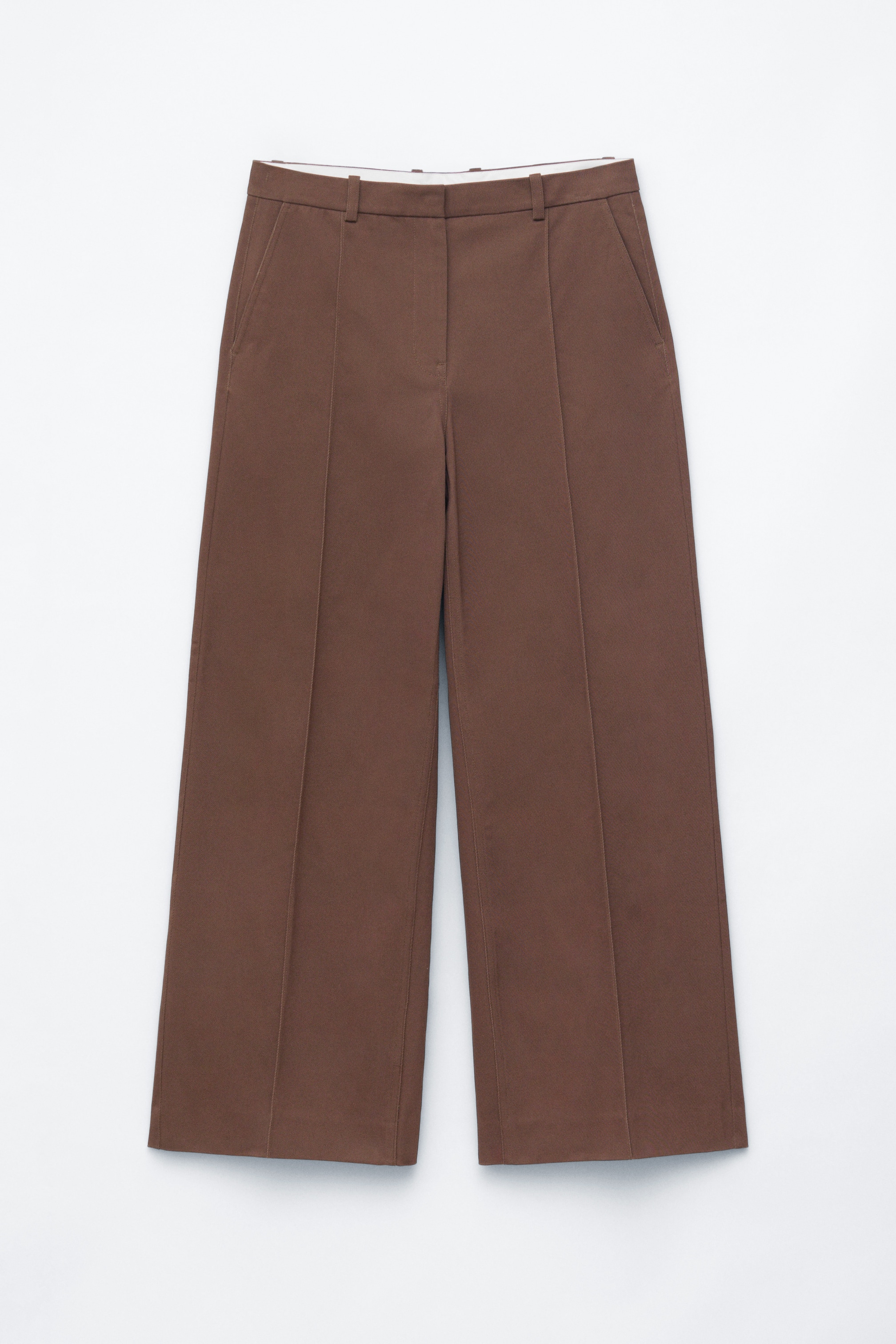 Ver imagem maior: Calças castanhas apresentam uma vista frontal, com um cós liso com passadeiras para cinto e bolsos laterais inclinados nas costuras. São desenhadas com pernas largas e vincos acentuados que percorrem o centro frontal de cada perna.