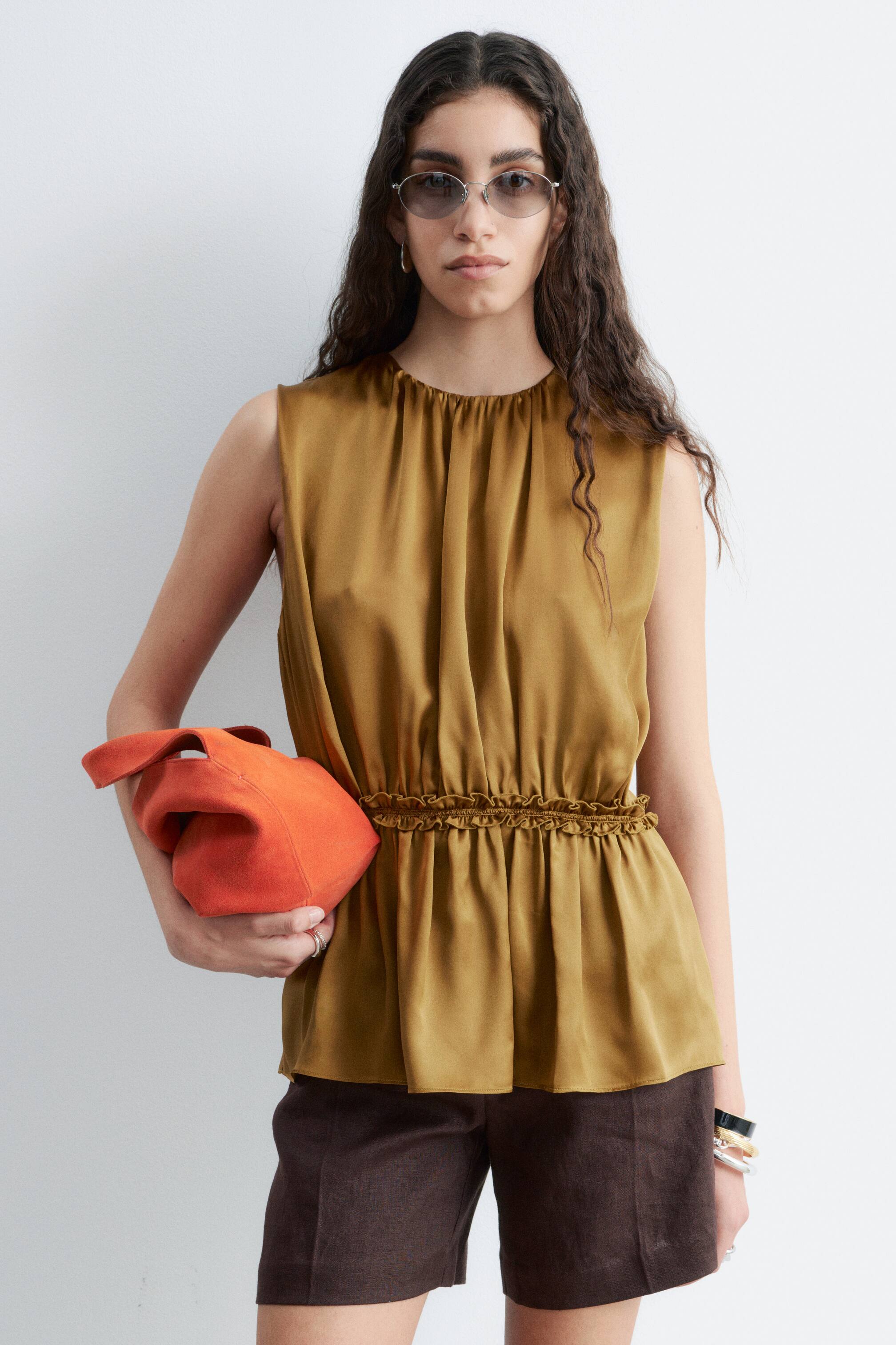 Visa större bild: En skimrande brons-guld ärmlös topp i ett satinliknande tyg med en rynkad rund halsringning och en åtsittande elastisk midja med volangdetaljer, vilket skapar en peplumfåll.