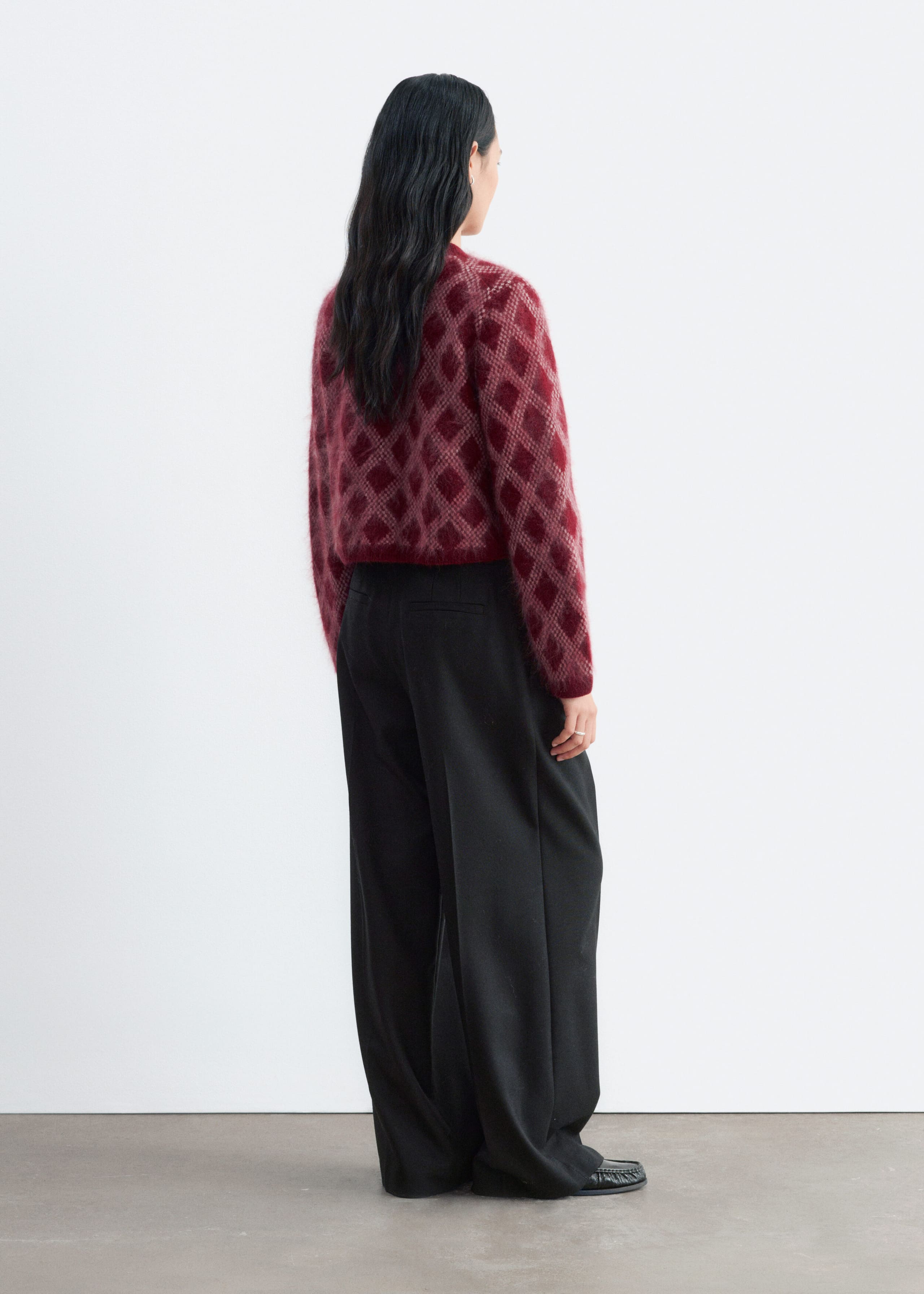 A fuzzy knit cardigan with a red and pink diamond pattern is being worn by the model, styled with black wide-leg trousers. The cardigan features a cropped silhouette, structured shoulders, and ribbed trims visible from the back.