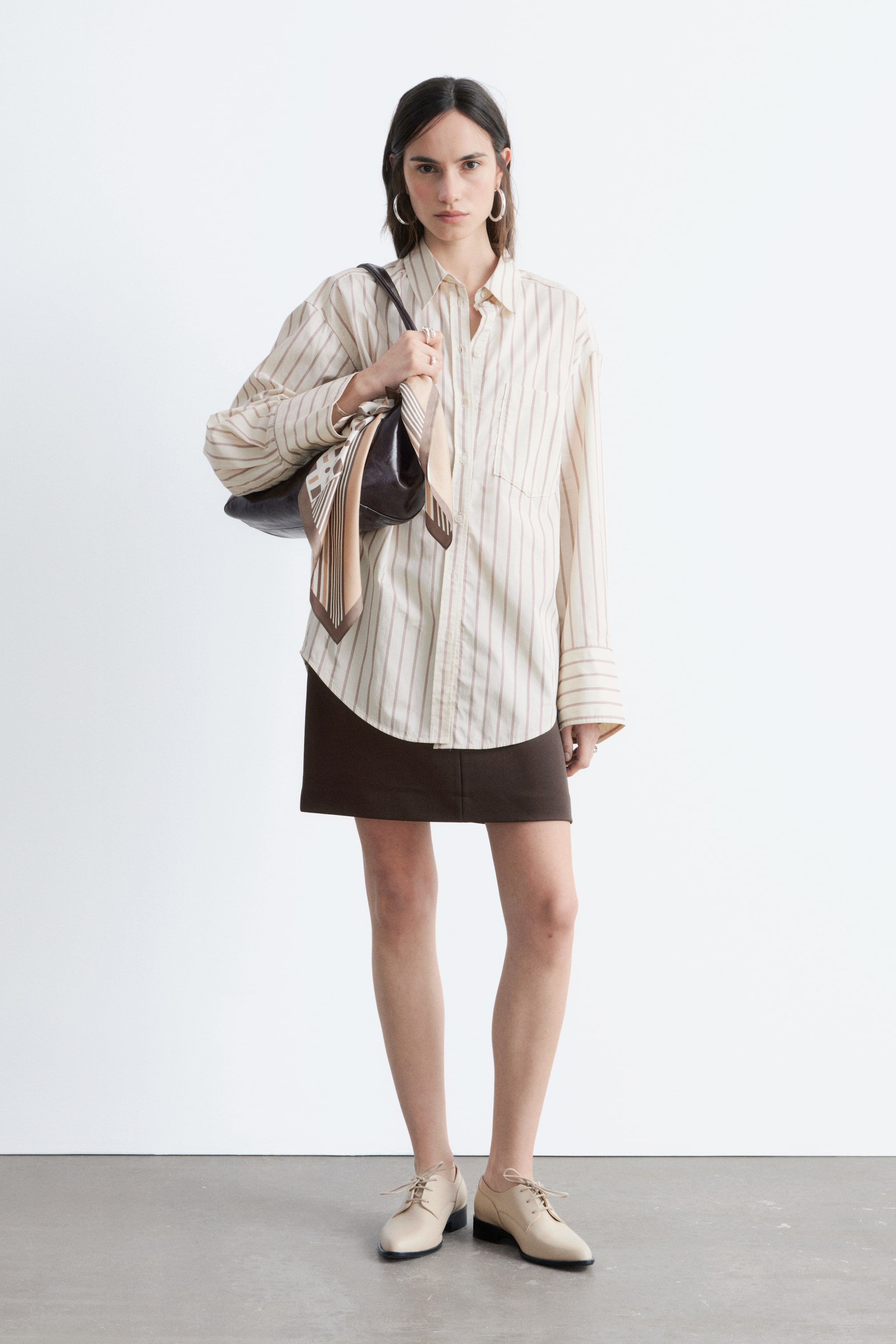 Visualizza immagine più grande: La camicia color crema con bottoni presenta sottili righe verticali marroni, un colletto a punta, una chiusura frontale con bottoni e una tasca applicata sul petto sinistro. Si distingue per una vestibilità oversize con maniche lunghe e ampie e polsini estesi, indossata frontalmente con l'orlo arrotondato che cade fuori da una minigonna marrone scuro.